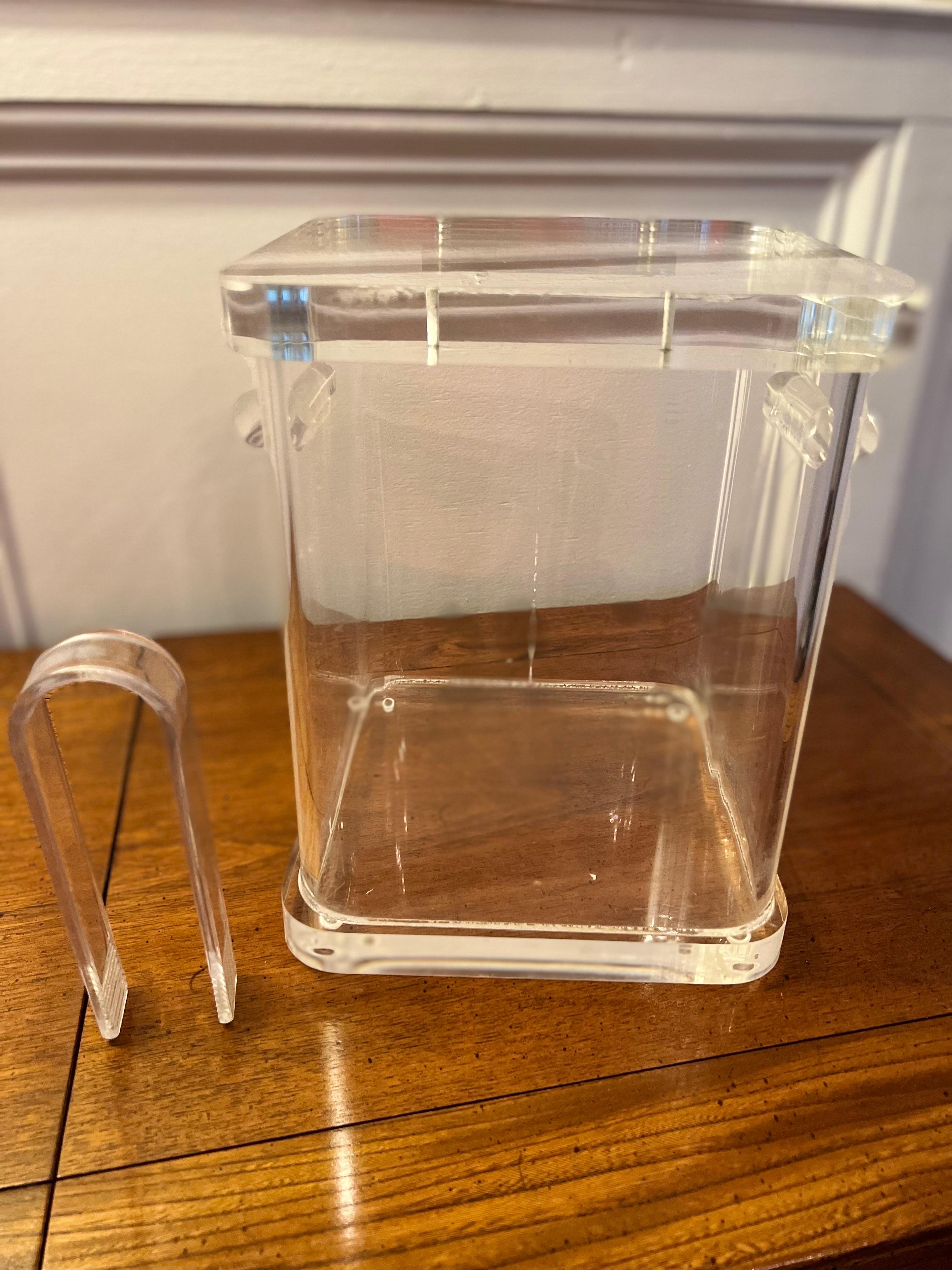 1970's vintage chunky lucite ice bucket with solid cylinder handles, tongs and hinged lid. The bucket has a rounded square shape and is well crafted having good weight to it. The lucite is in nice condition. 3/4