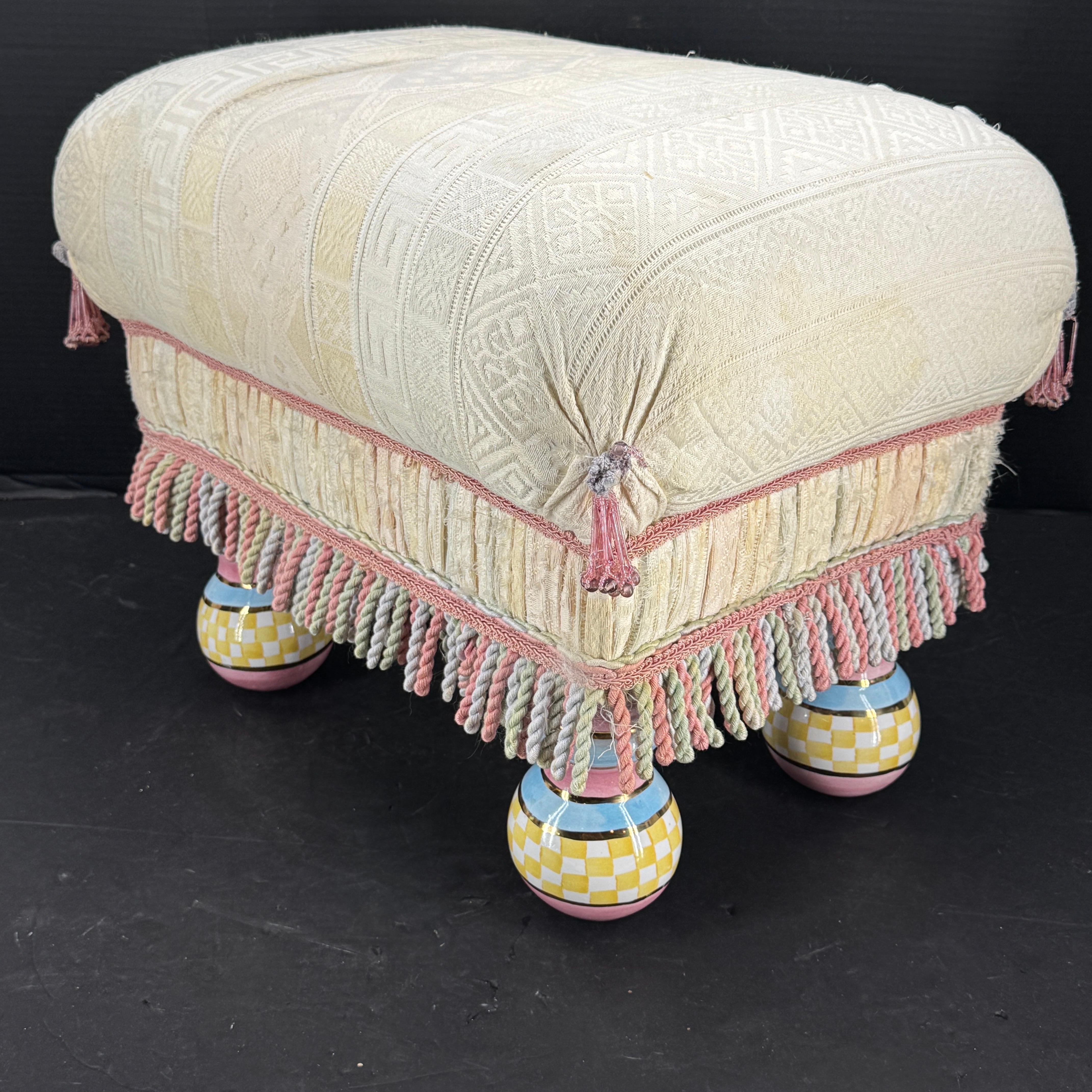 Stylish and classic best describe this vintage upholstered footstool from Mackenzie Childs. The eye catching hand-painted majolica with ceramic feet certainly make a statement!  Adorned with a neutral fabric with tassels and fringe, this piece is