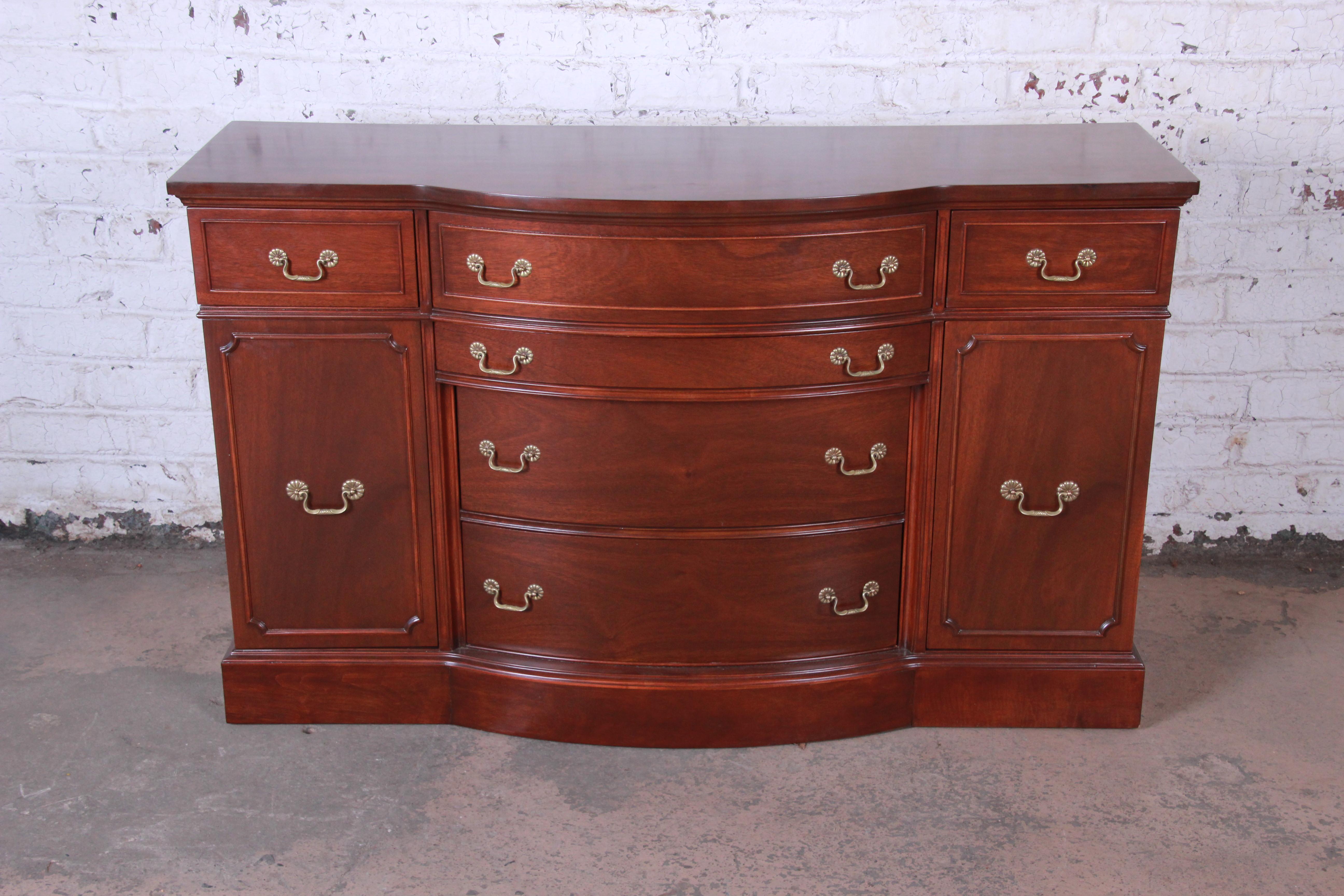 A gorgeous vintage mahogany bow front sideboard buffet. The sideboard features stunning mahogany wood grain and stylish original brass hardware. It offers ample room for storage, with six deep drawers and two cabinets. The sideboard has recently
