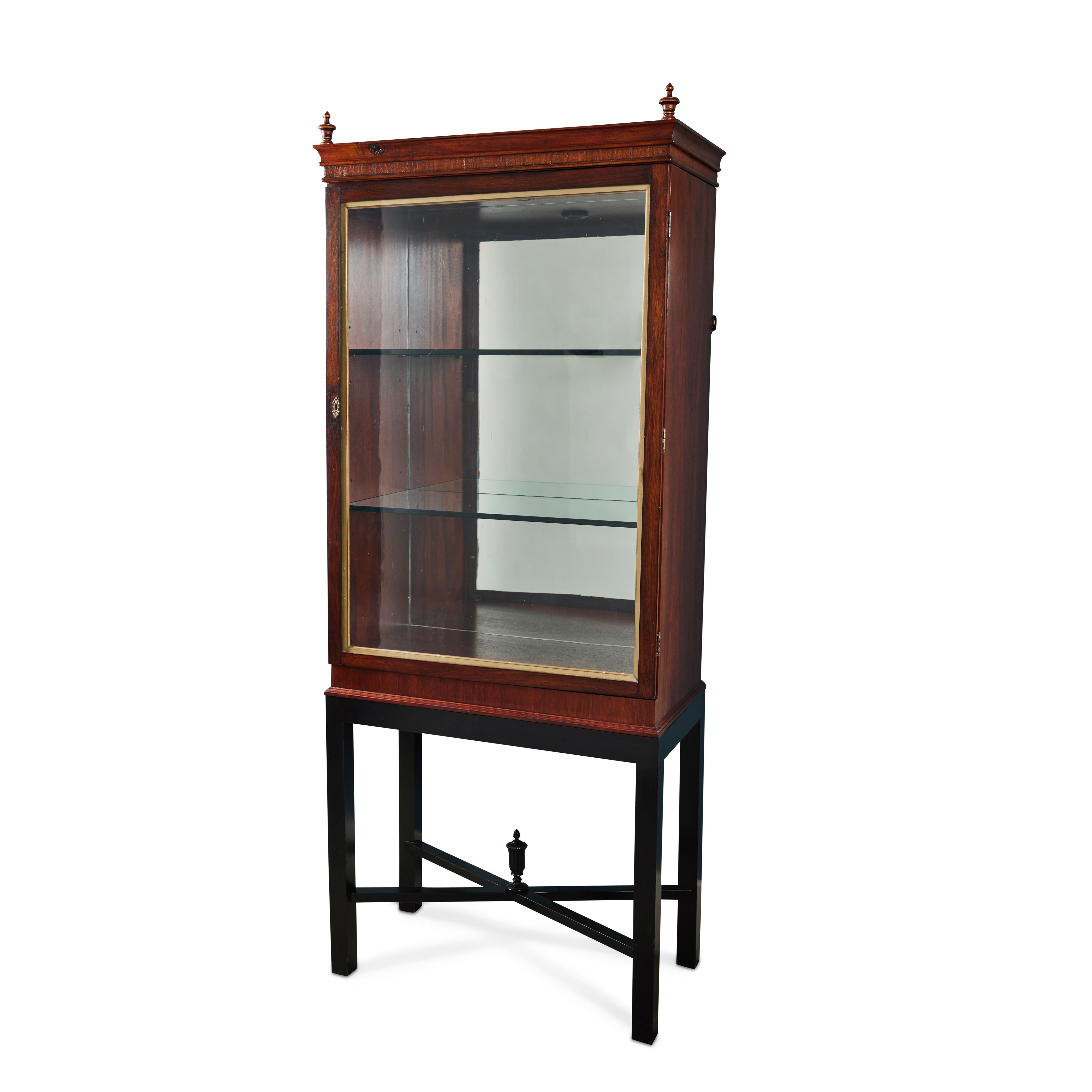 This elegant vintage mahogany display cabinet has a mirrored back and two glass shelves to exhibit your special treasures.  Adorned with black cross-bar connecting legs and a decorative urn detail, it comes with a gorgeous hand blown glass door and