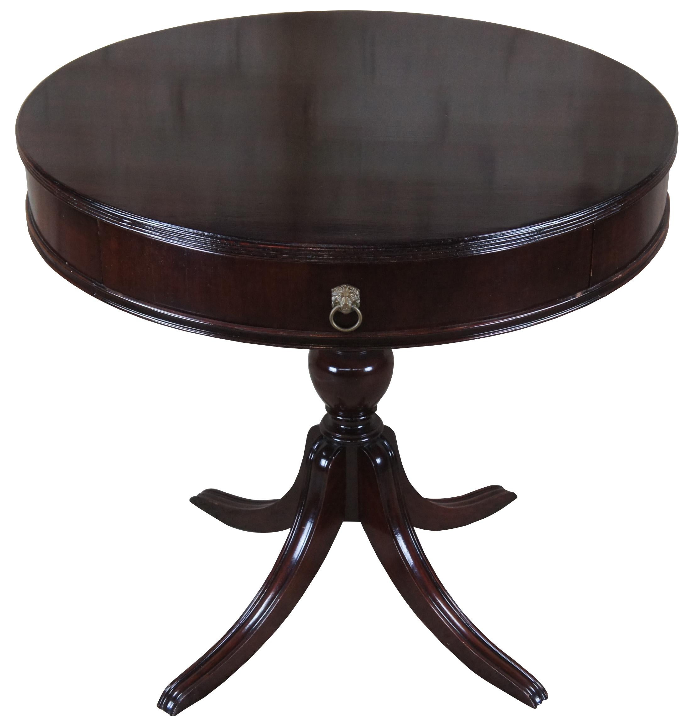 Sheraton style mid 20th century mahogany drum table with glass top. Features a round top with fluted edge and two drawers over a turned urn shaped baluster base leading to four saber legs. Includes gold lion head knockers along the drawers. A great