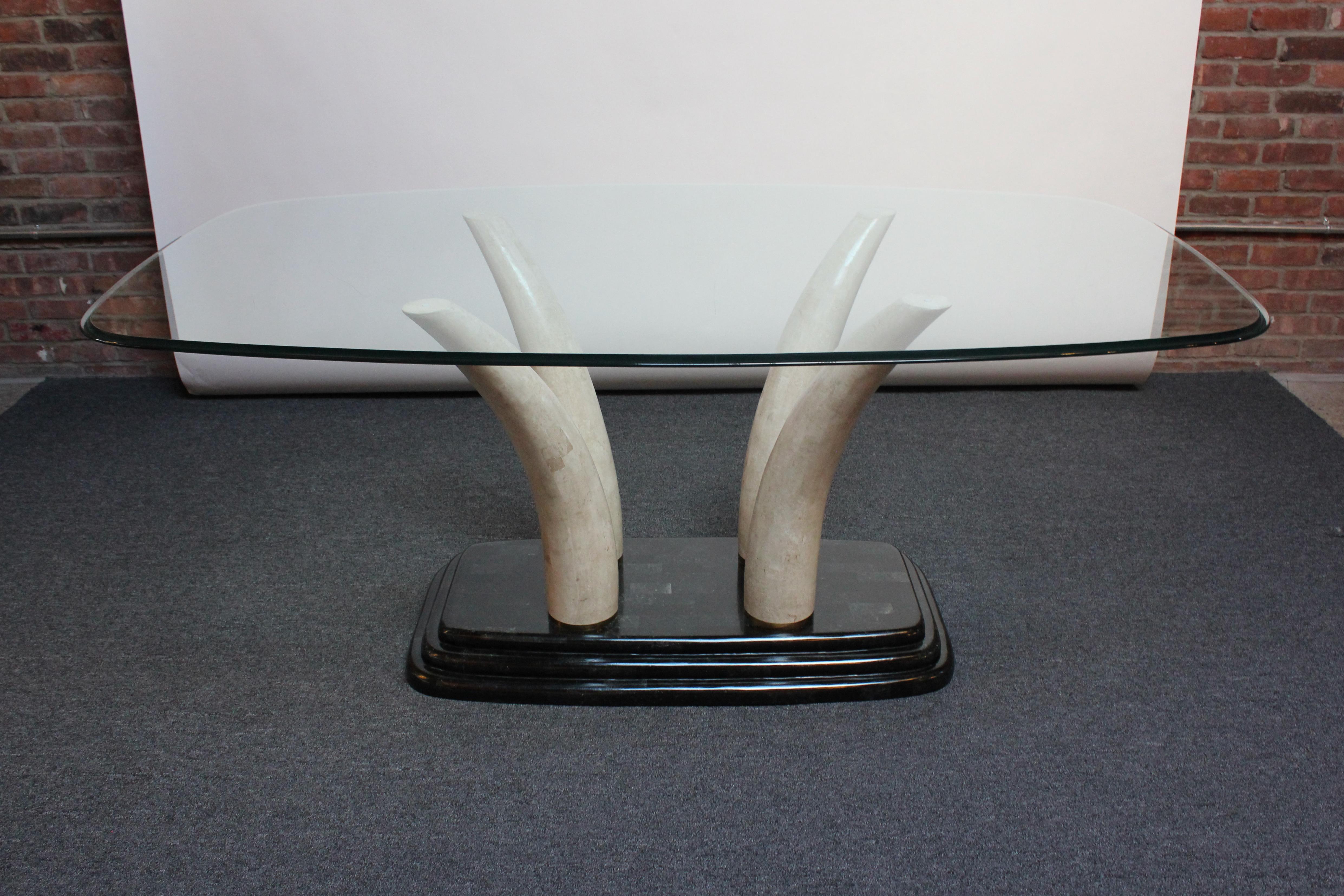 Maitland Smith dining table composed of a tempered glass top with Ogee edge supported by four splayed faux-elephant tusks in tessellated stone / travertine connected by brass fittings to a heavy stepped marble base in ebony with green tint.
The