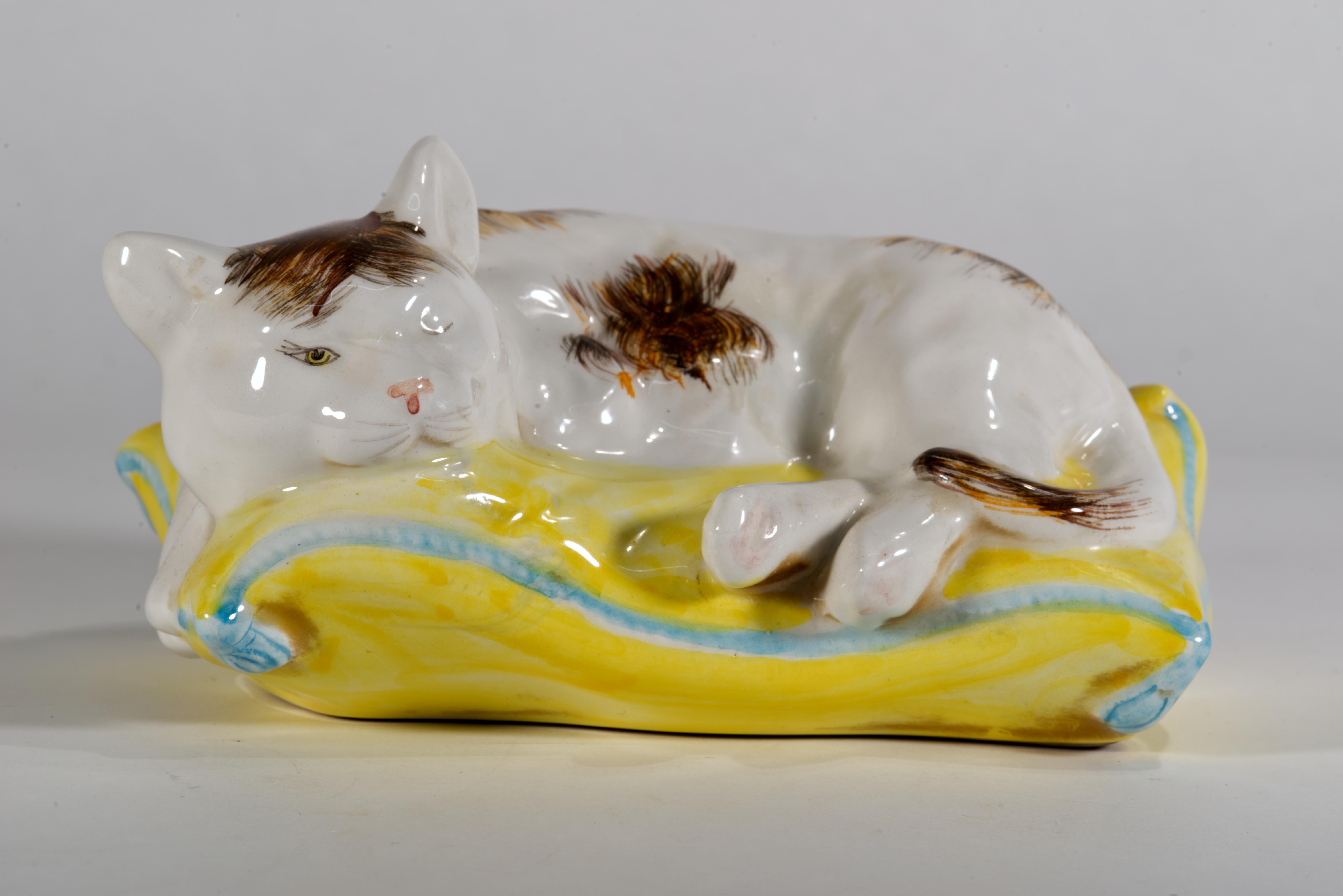 La scultura in ceramica vintage raffigura un gatto o un gattino soriano che riposa sul cuscino con la testa leggermente sollevata e girata di lato. La statuetta è decorata con uno smalto di maiolica con segni dipinti a mano di colore grigio scuro,