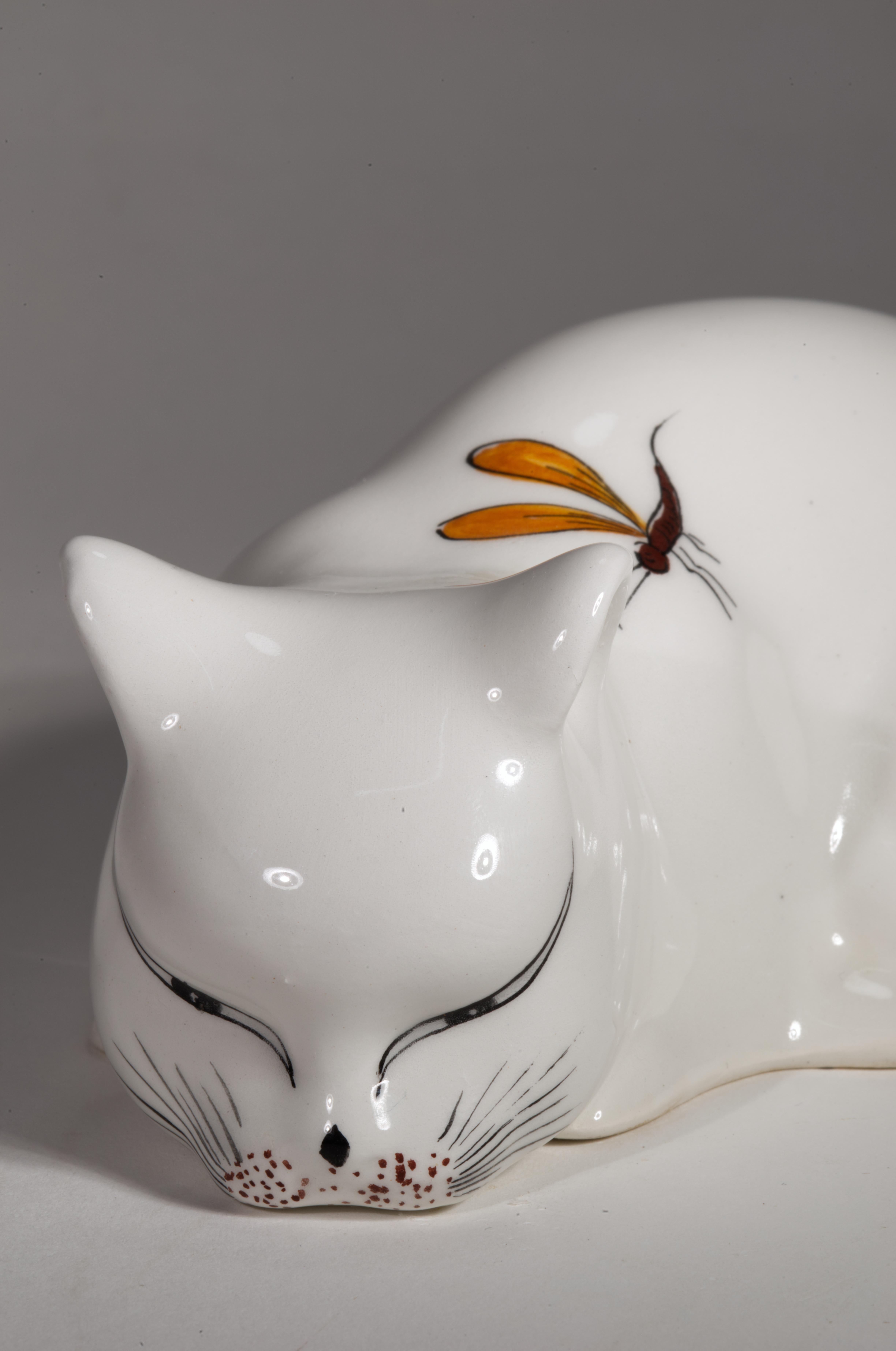 Playful Italian pottery cat figurine features crouching white cat with half-closed eyes, getting ready to pounce on a dragonfly flying over cat's shoulder. Elegantly sculpted ceramic cat body is finished with glossy white majolica glaze, and cat's
