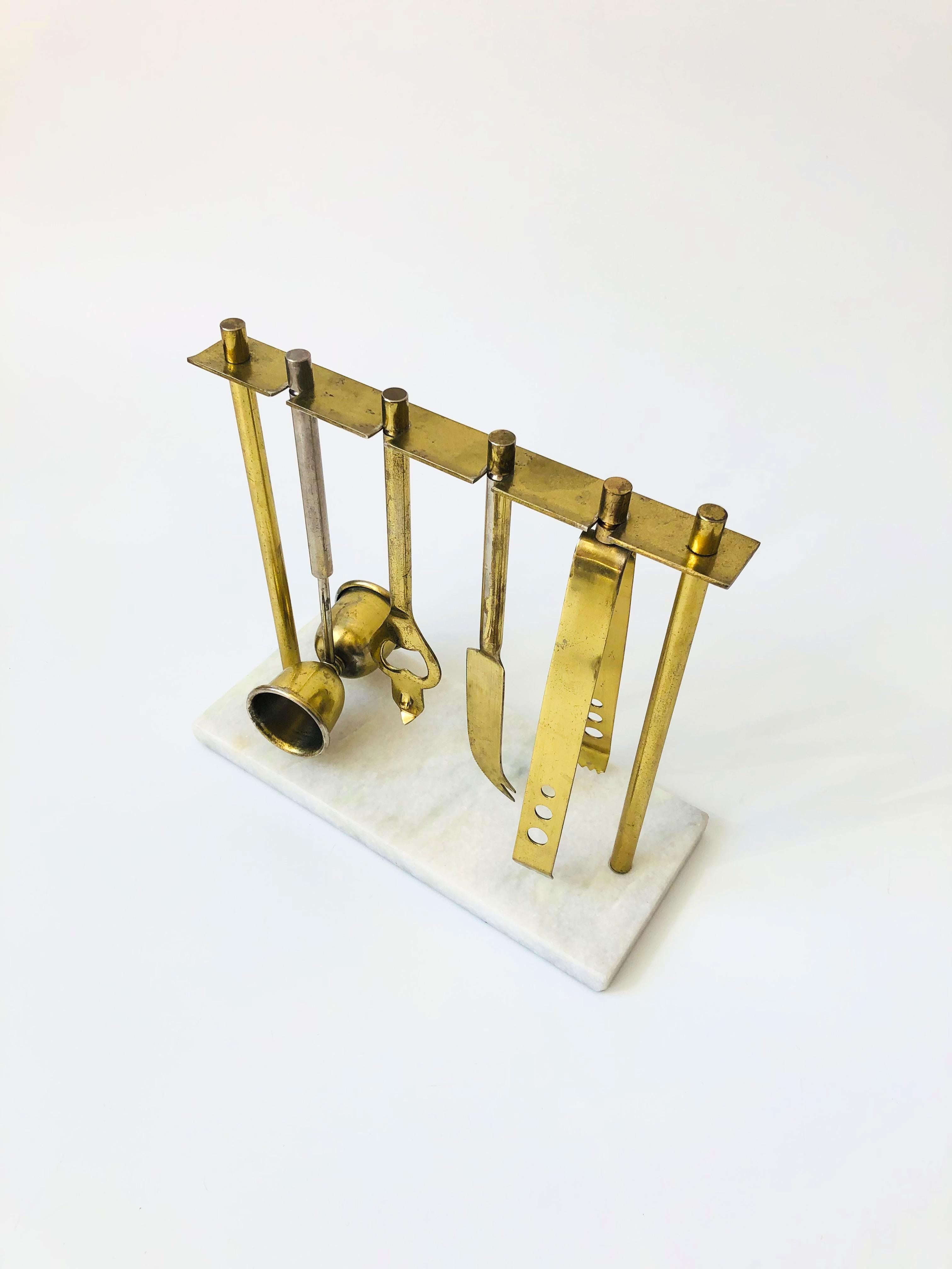 A vintage brass bar tool set with a hanging stand. Each piece is made of brass, hanging on a brass frame that is mounted to a marble base. The set includes ice tongs, a cocktail knife, a two sided jigger, and a bottle opener.
   