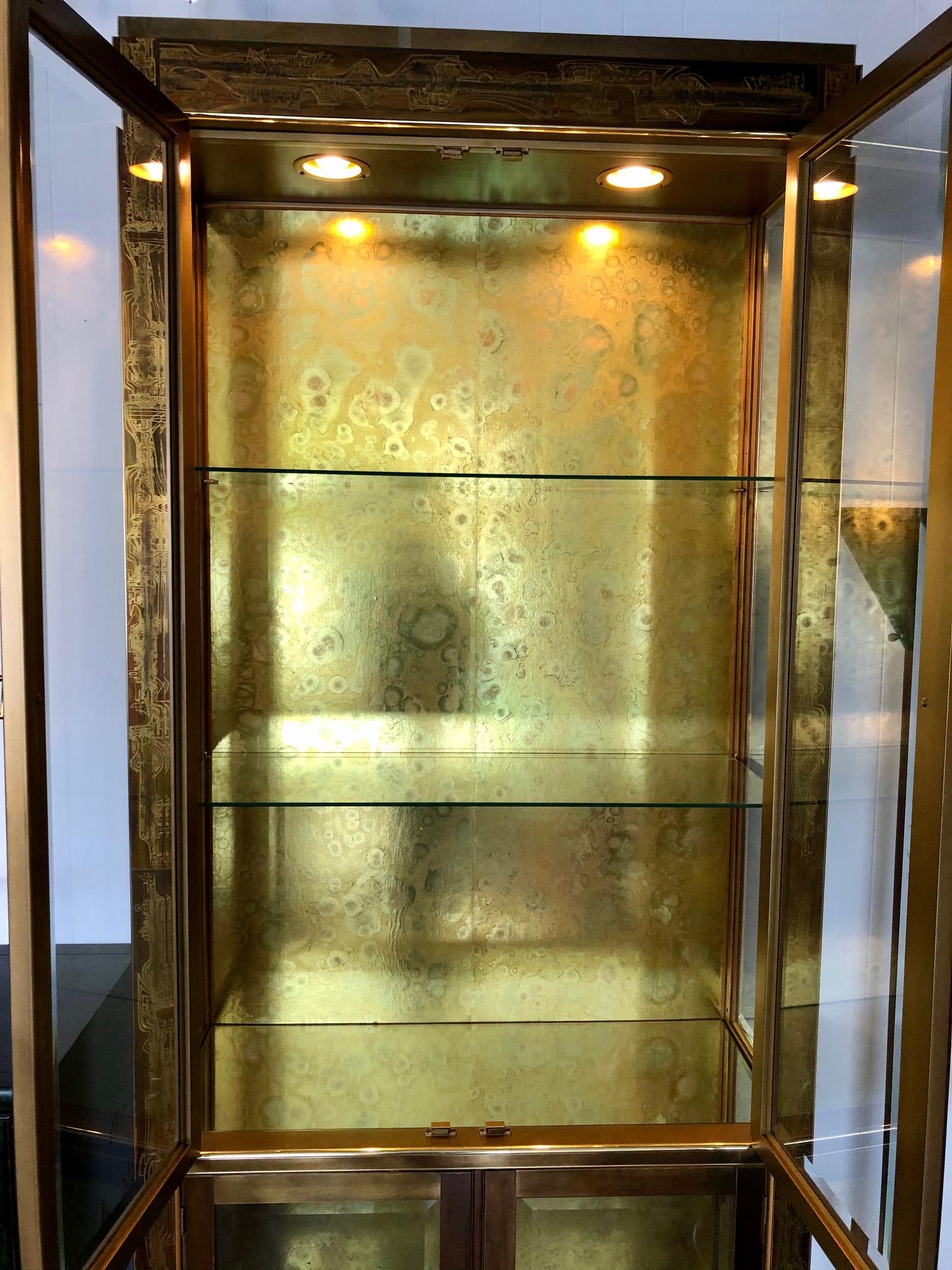 This pair of brass and glass vitrines by Mastercraft is in overall good condition. Designed by Bernhard Rohne. Acid-etched. Labeled in back. Four beveled glass front panels doors. Gold print interior. Glass shelves. Mirror shelves. Two lights, top