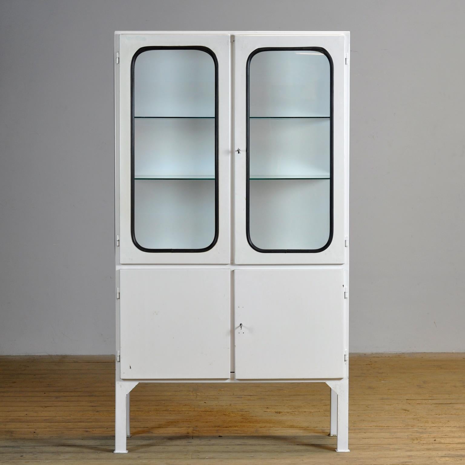 Ce cabinet médical a été conçu dans les années 1970 et a été produit vers 1975 en Hongrie. Elle est faite de fer et de verre avec de nouvelles étagères en verre. Le verre est maintenu par une bande de caoutchouc noir. L'armoire comporte deux