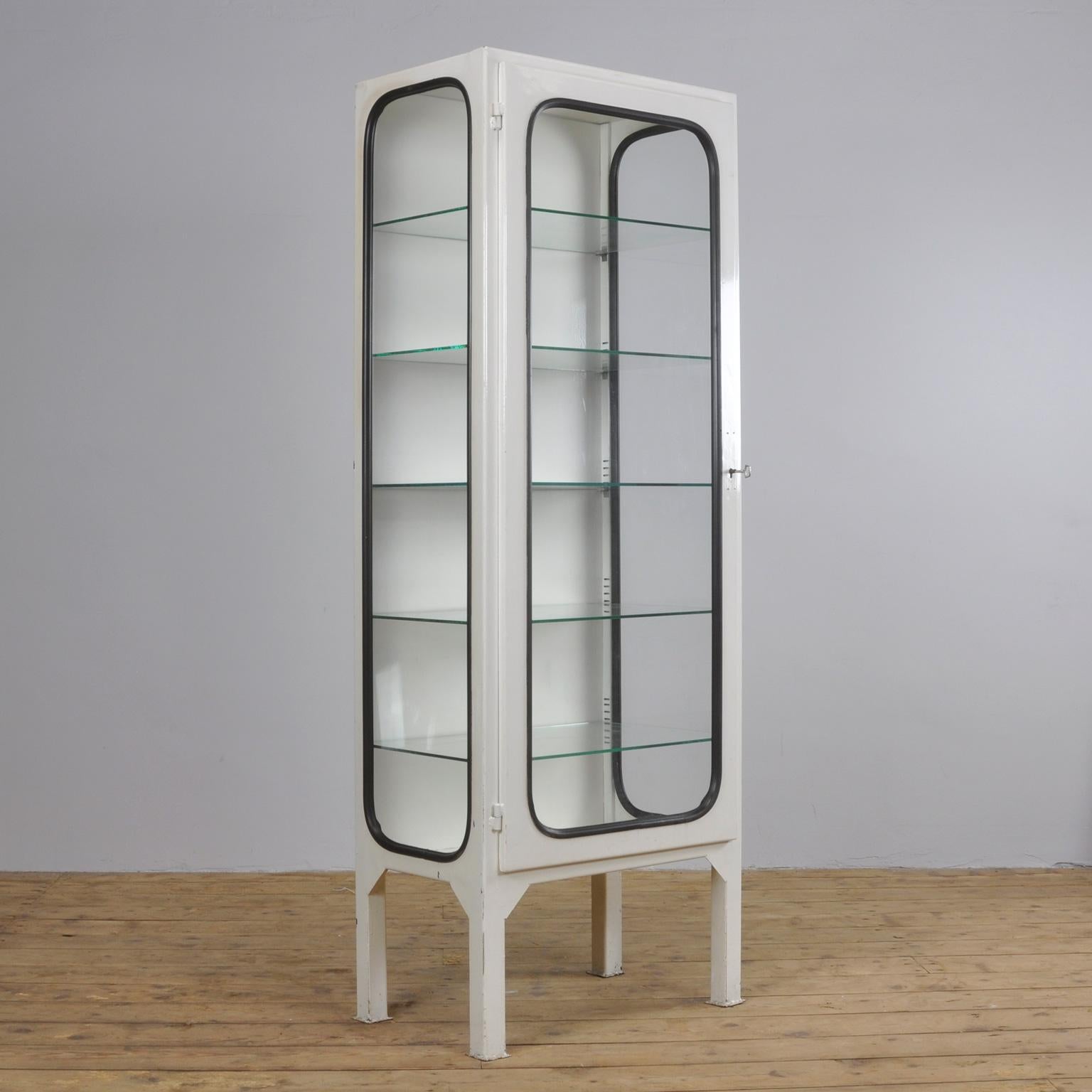 This medicine cabinet was designed in the 1970s and produced in 1975 in Hungary. It is made from steel and glass, with the glass panes held in place by a black rubber strip. The cabinet features five adjustable glass shelves and a functioning lock.