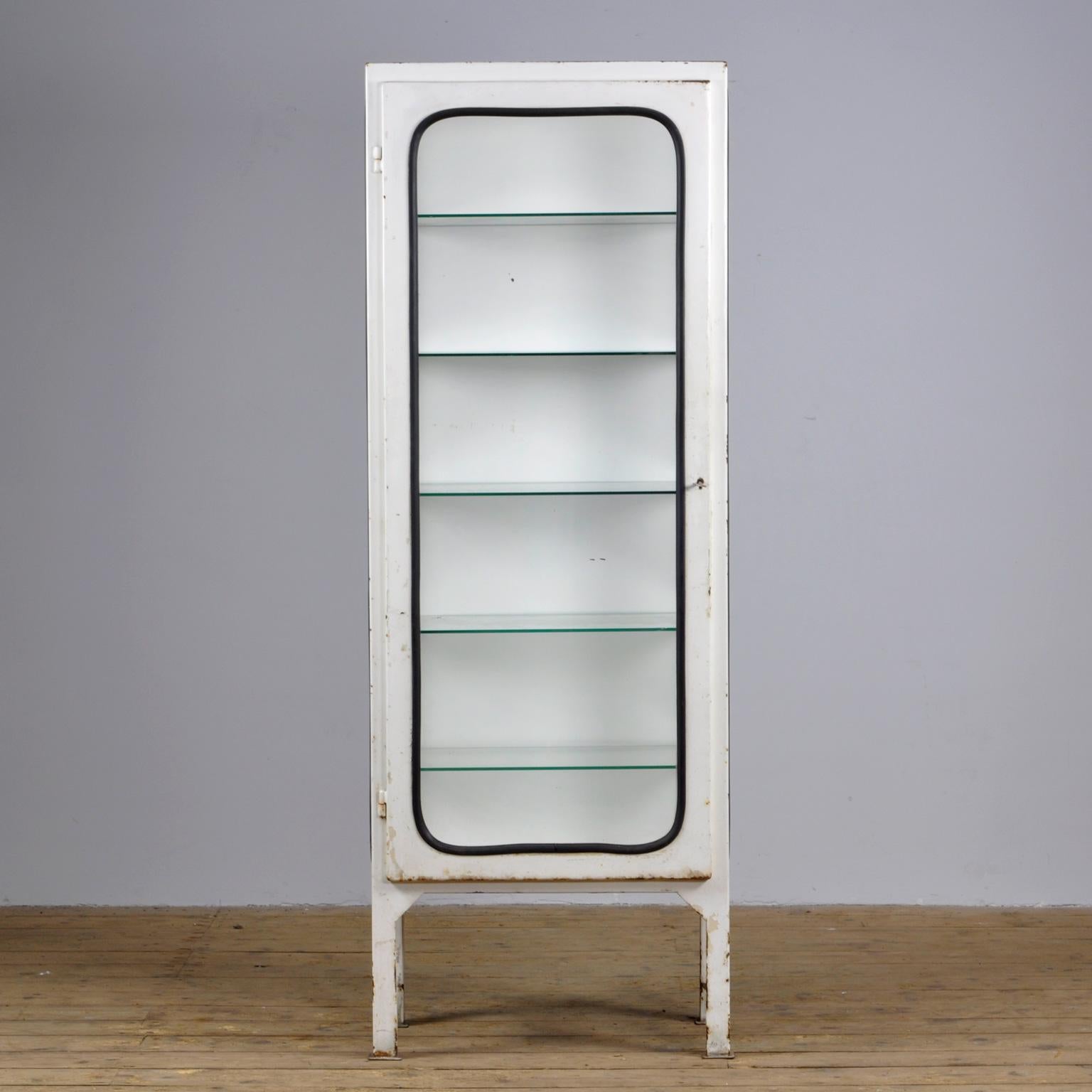 This medicine cabinet was designed in the 1970s and produced in 1975 in Hungary. It is made from steel and glass, with the glass panes held in place by a black rubber strip. The cabinet features five adjustable glass shelves and a functioning lock.