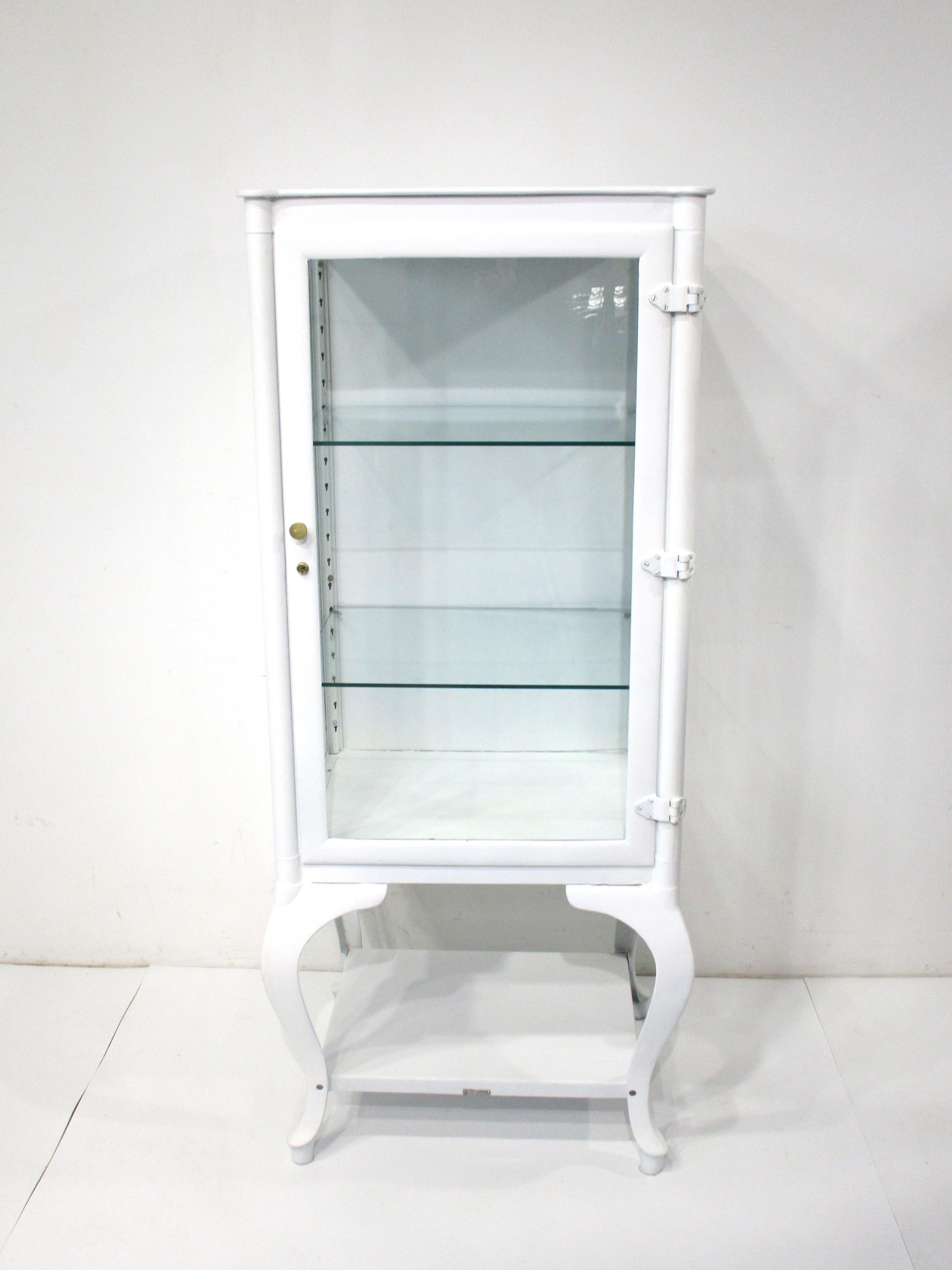 A heavy duty well made white painted metal medical cabinet with three sides in glass having two adjustable glass shelves and pins for a third shelve . Dated 1907 as the patented date with swing out door , brass hardware and lower storage area . The