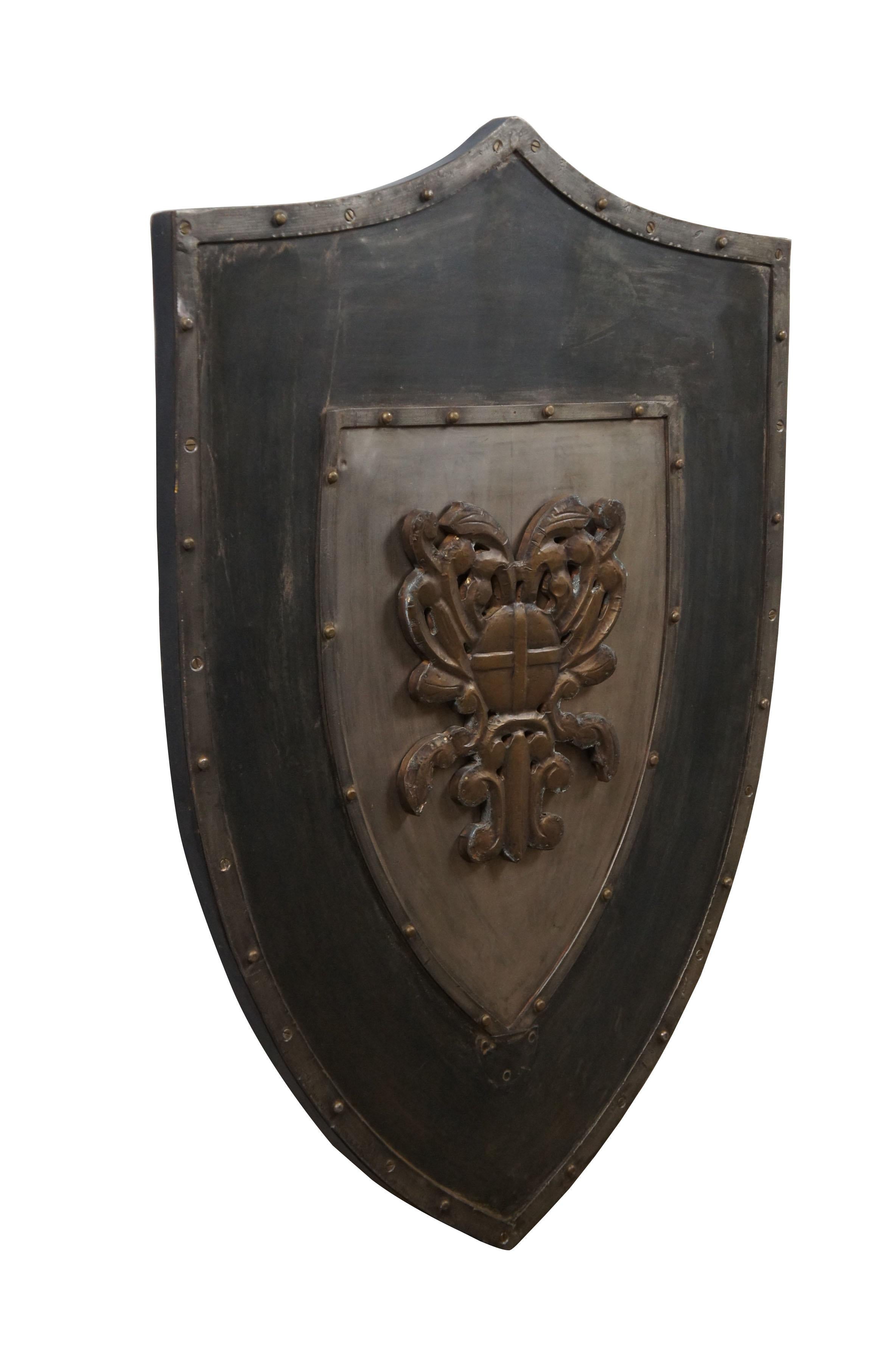 Late 20th century wall hanging shield - traditional shield shape crafted in dark patinaed metal on a wood base, with riveted boders around a central brass colored motif resembling a coat of arms with swirling folaite design around a royal orb. Two