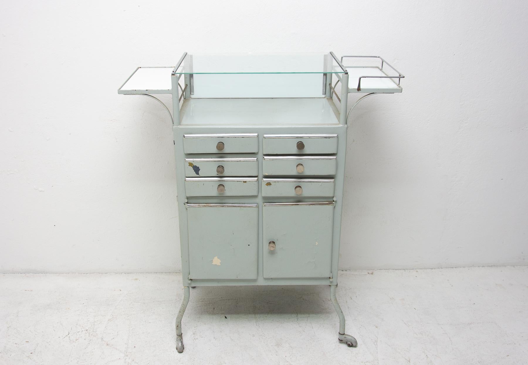 Metal and glass medical/apothecary cabinet on wheels with nice patina.
This cabinet was made in the former Czechoslovakia in the 1960s.
One upper glass shelves, two on the sides, six drawers and bottom storage space
Original paint, in good vintage