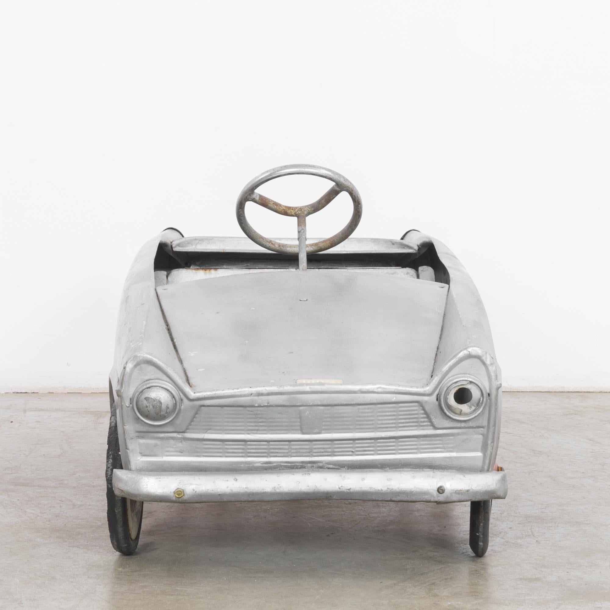 A metal pedal car from the 1960s. This sophisticated toy has the long, low, flat shape of a vintage car; a raised steering wheel and the back license plate add to this charming impression. The body of the car is painted a matte silver-gray, while