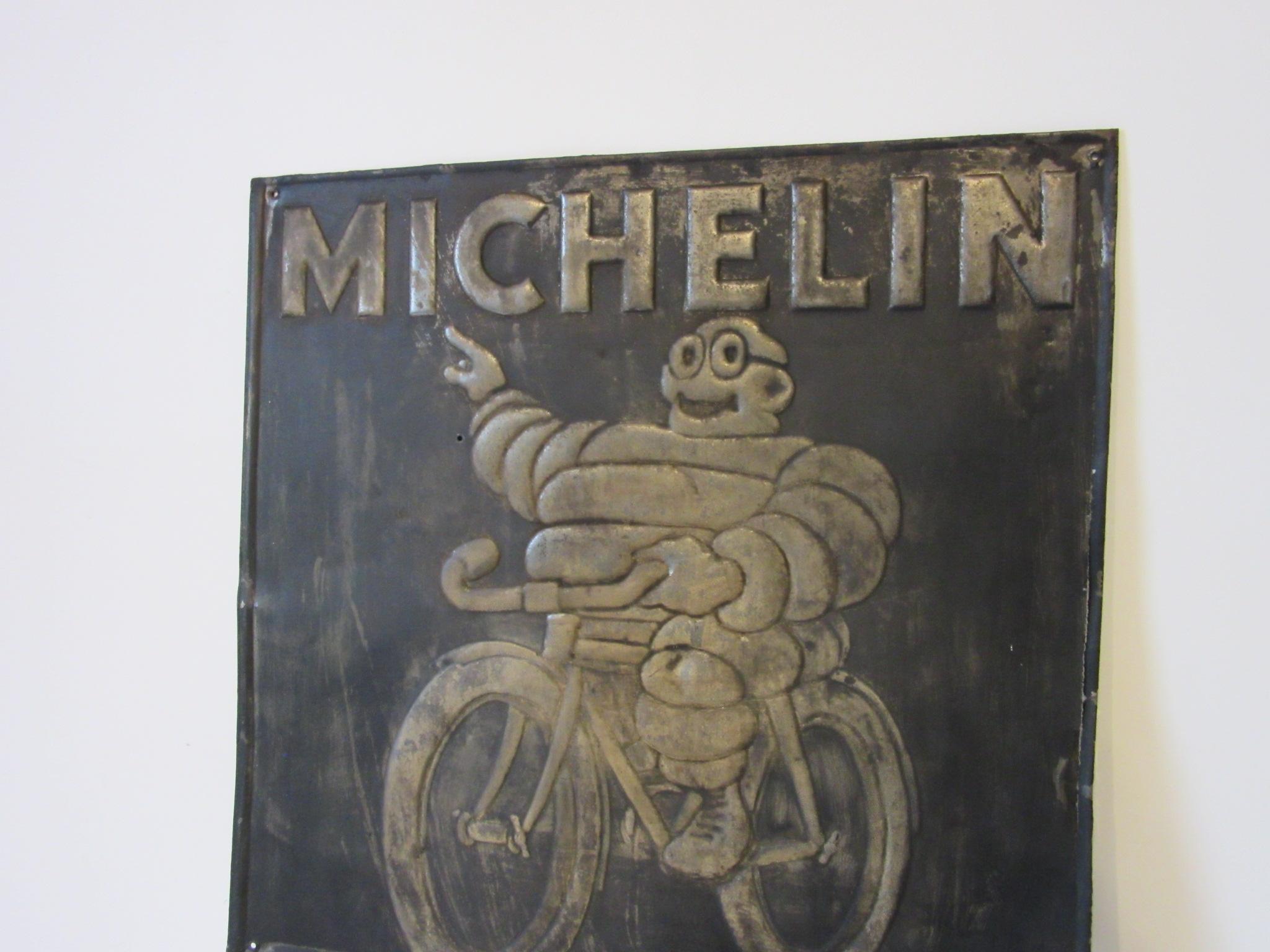 A vintage embossed metal Michelin tire advertising sign depicting the ever famous Bibendum or Michelin Tire man who is the mascot for the company and the worlds oldest trademark. Bibendum is pictured riding a bicycle and promoting the tire brand,