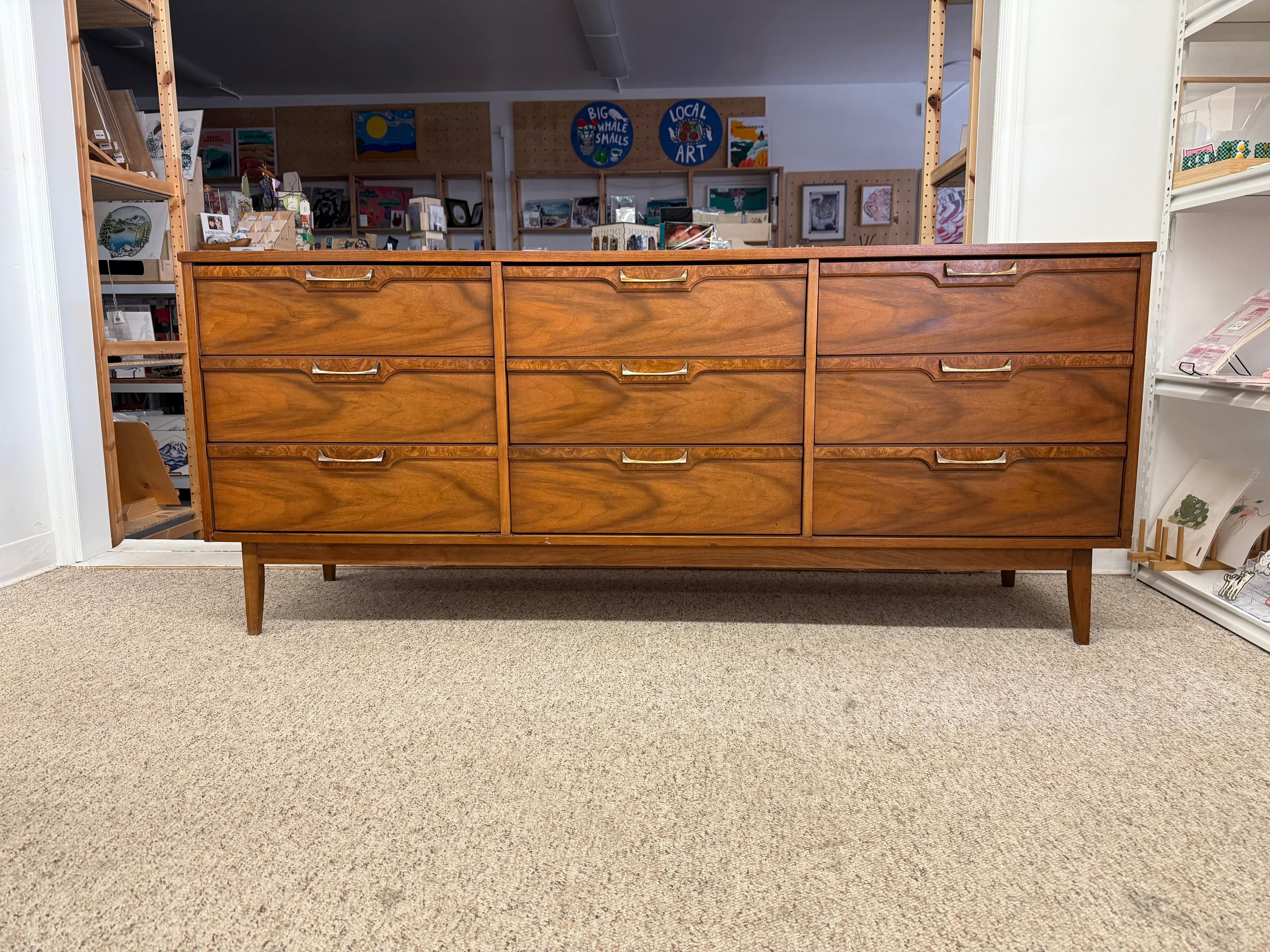 The dresser is a mid-century modern piece with a long, low-profile design, featuring nine drawers in a 3x3 configuration. Crafted from wood with a warm, medium-brown finish, likely walnut or a similar hardwood, it showcases prominent grain patterns