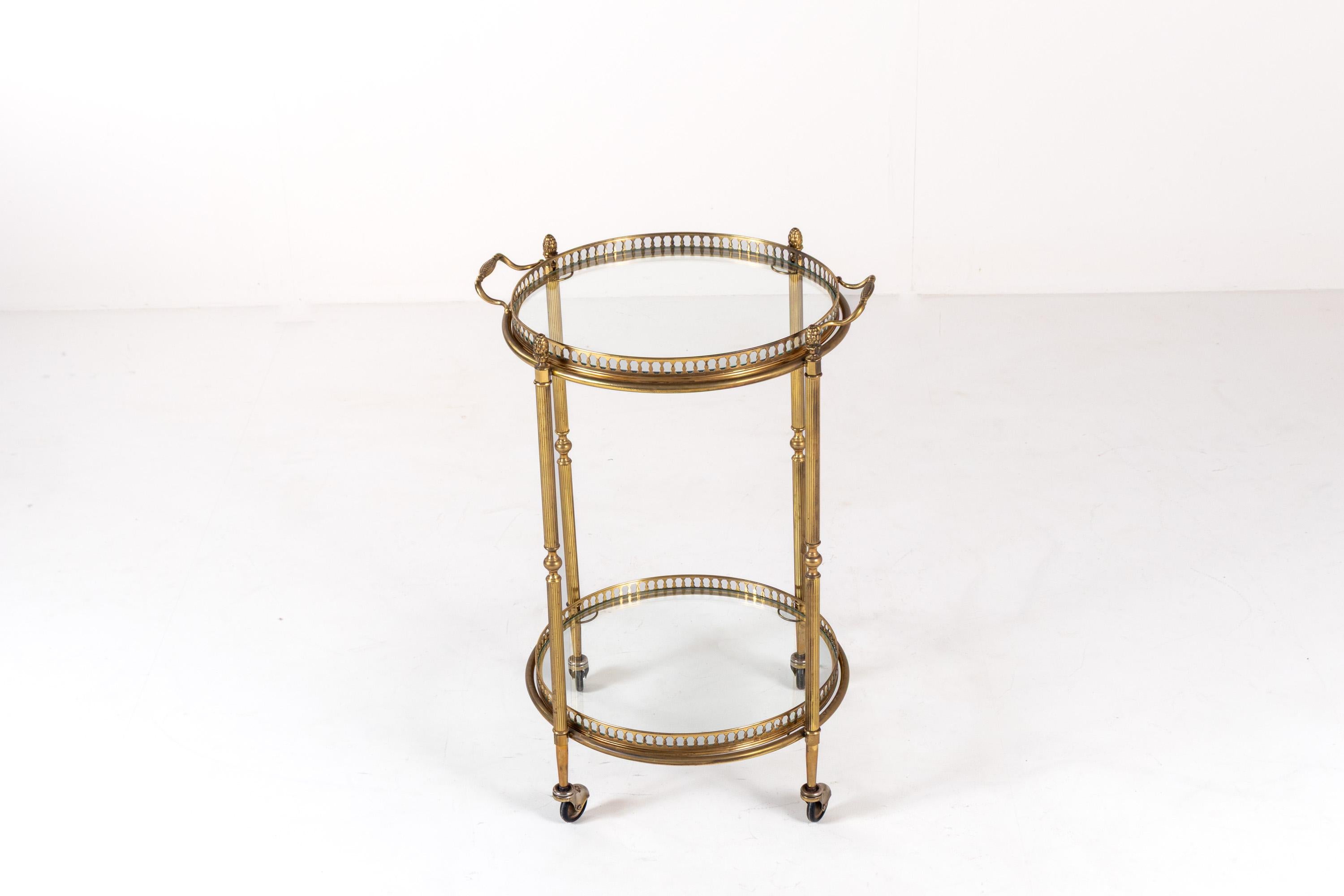 This exquisite vintage brass bar cart exudes elegance and sophistication, featuring a two-tier design with glass shelves and intricate detailing. The circular structure is supported by four finely crafted fluted legs, adorned with delicate accents.