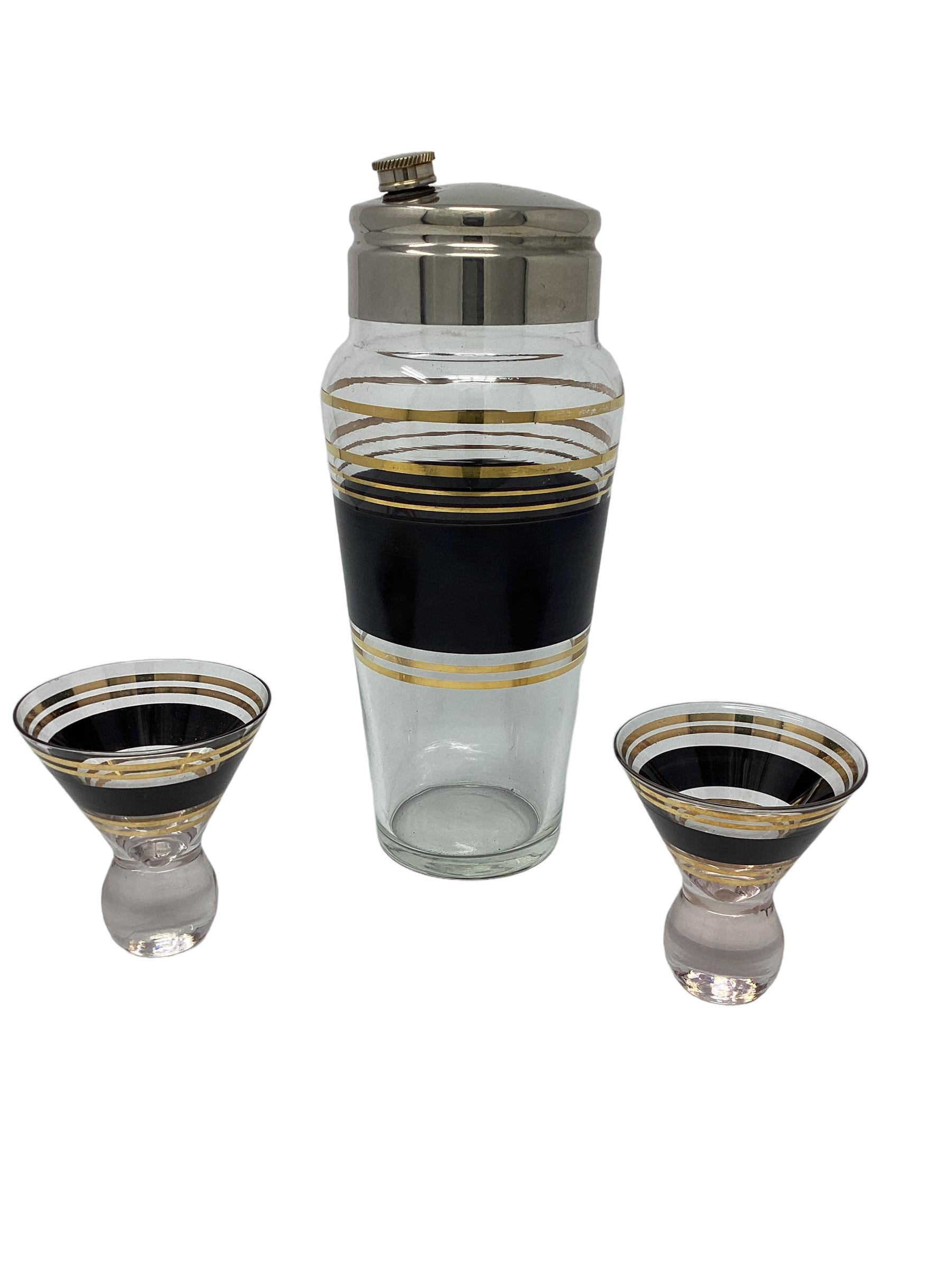 Vintage Mid Century Cocktail Set decorated with a wide band in black enamel  and gold bands. The shaker is topped with a chrome cover. The cocktail cups have a round weighted base and measure 3.125”w x 3.5”h.