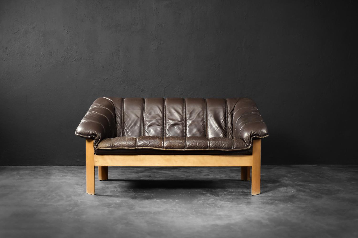 This two-seater sofa was manufactured in Denmark during the 1970s. The frame is crafted from solid wood in a honey-brown shade. It is upholstered in dark chocolate-colored genuine leather. The soft, tufted seat with wide armrests is set within the