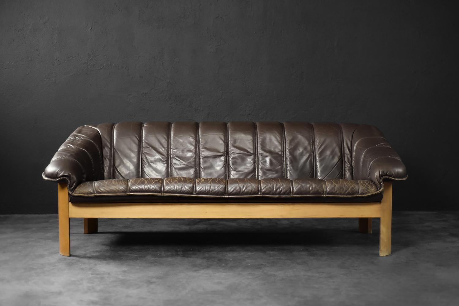 This three-seater sofa was manufactured in Denmark during the 1970s. The frame is crafted from solid wood in a honey-brown shade. It is upholstered in dark chocolate-colored genuine leather. The soft, tufted seat with wide armrests is set within the