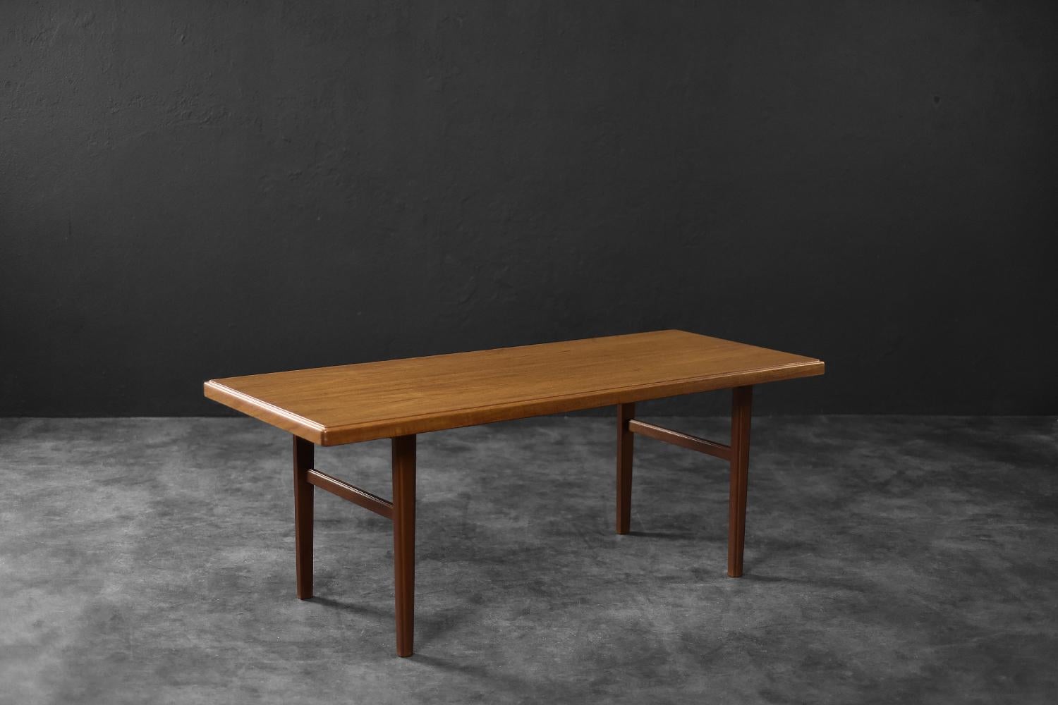 Cette longue table basse a été fabriquée au Danemark dans les années 1960. Le plateau est fabriqué en teck dans une teinte brun doré, avec un motif de grain clairement visible. La base comporte des pieds dans une configuration distinctive en forme