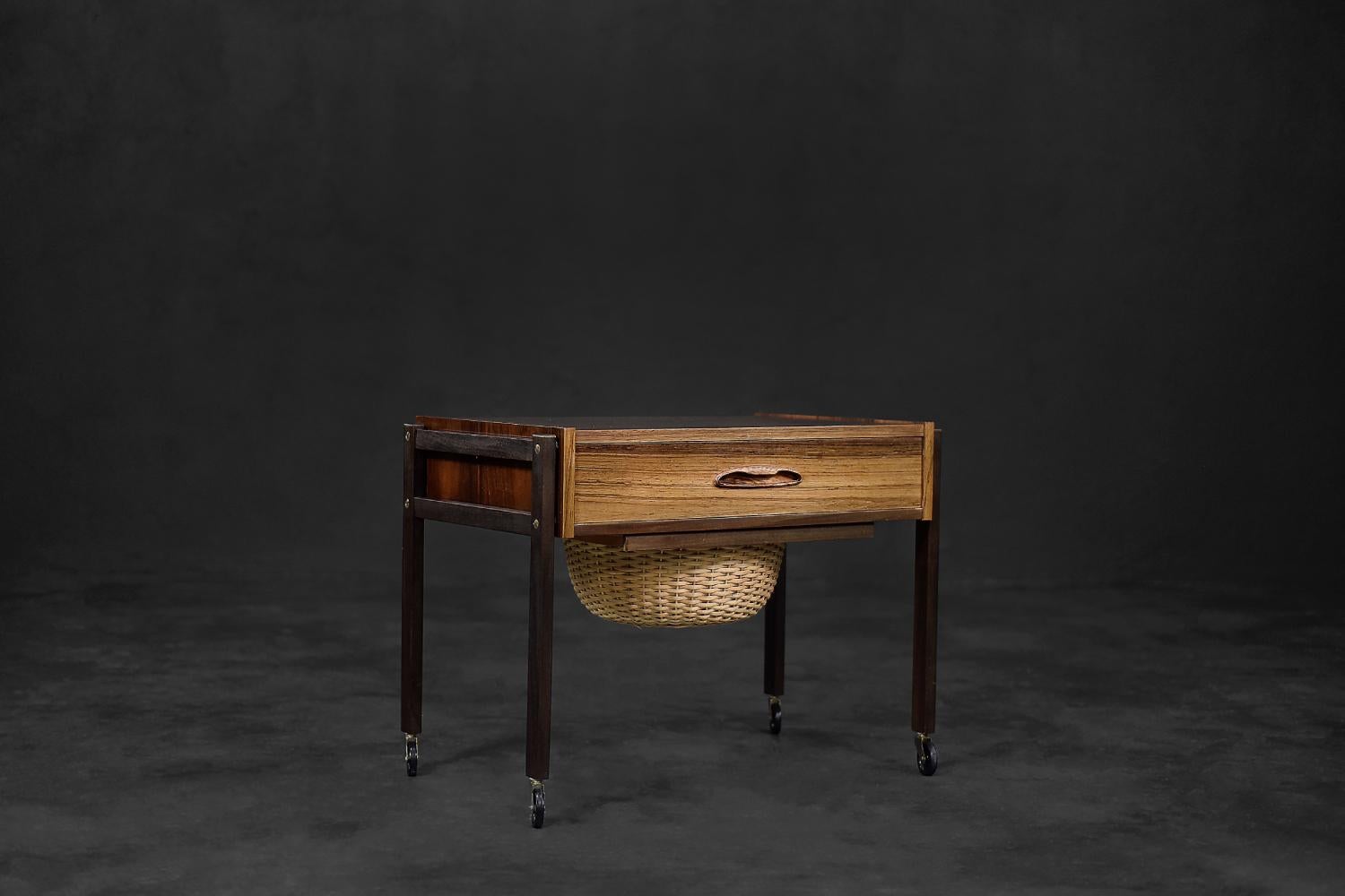 This mobile thread table was made in Denmark during the 1960s. It is made of rosewood in a dark shade of brown. It has a drawer with four compartments and a pull-out wicker basket. The legs are finished with small wheels, which allows for free