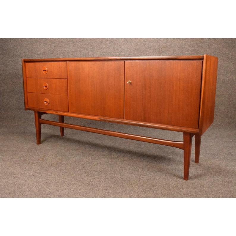 Here is a beautiful teak sideboard circa 1960 manufactured by Bartels In Germany.
This compact credenza, recently imported from Europe to California before its restoration, features a bank of three drawers and two large doors revealing a large