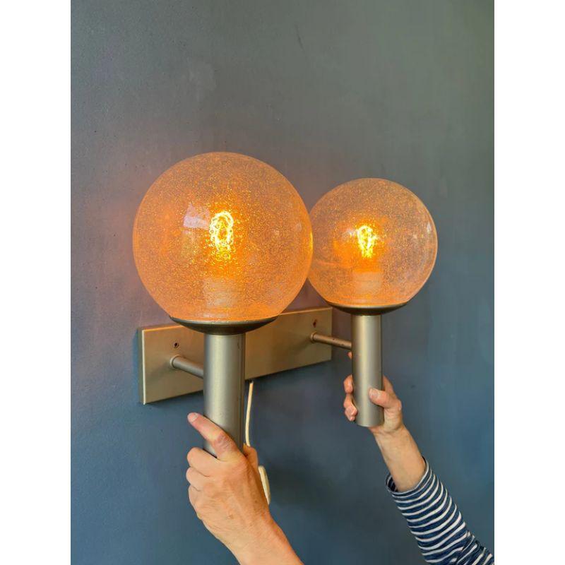 The double sconce wall light by Raak Amsterdam, Vigilante C1663. The wall lamp has a heavy metal frame and has two seeded glass shades. The lamp requires two E27 lightbulbs and currently has an EU-plug.

Dimensions:
ø Shades: 16 cm
Height: 29