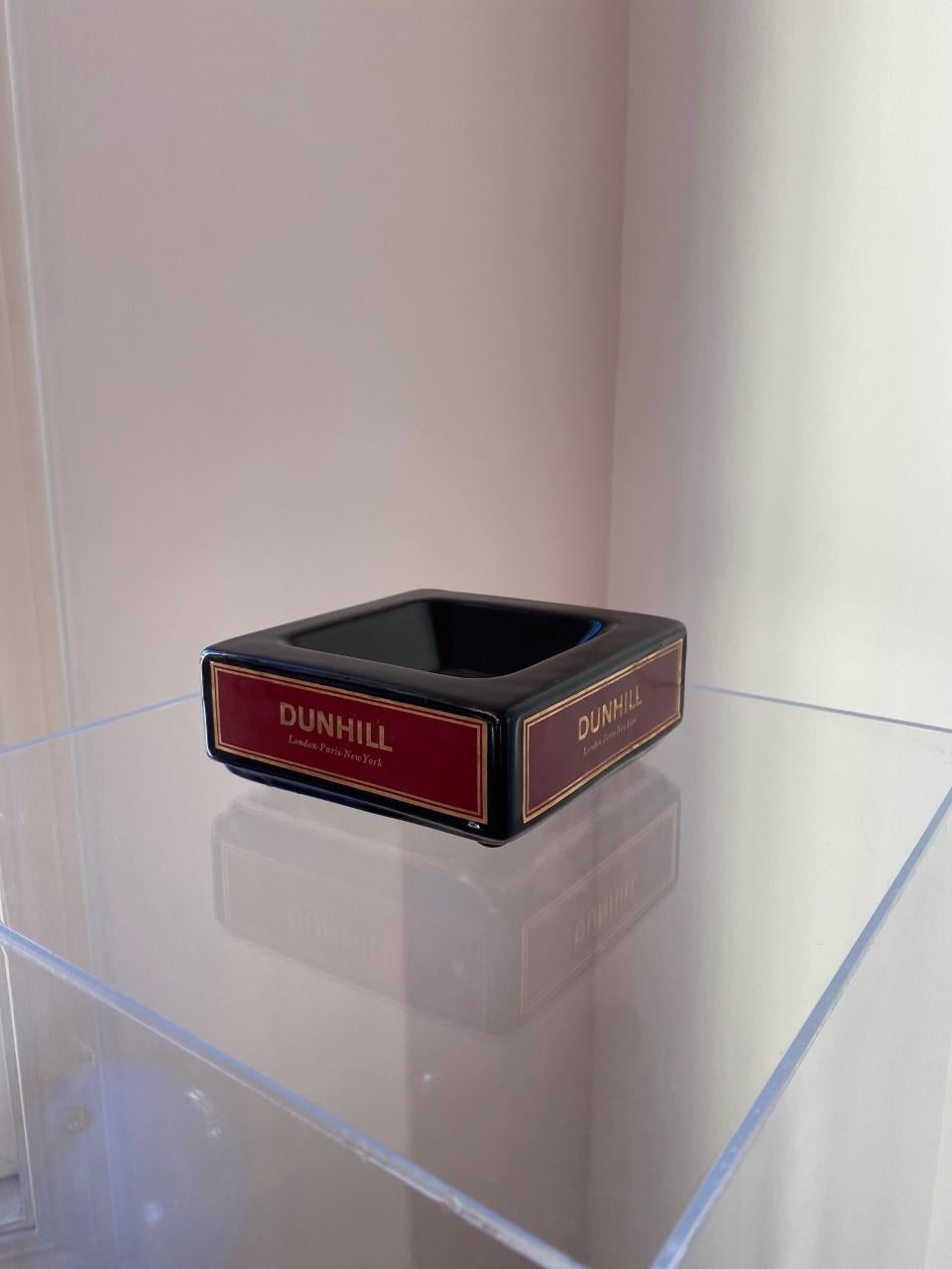 Square ceramic ashtray with black high gloss glaze, features red and gold Dunhill label on all four sides. Has four small feet to elevate it off a table surface. Dimensions: 5.75” width x 5.75” depth x 2.55” height. This piece is timeless and