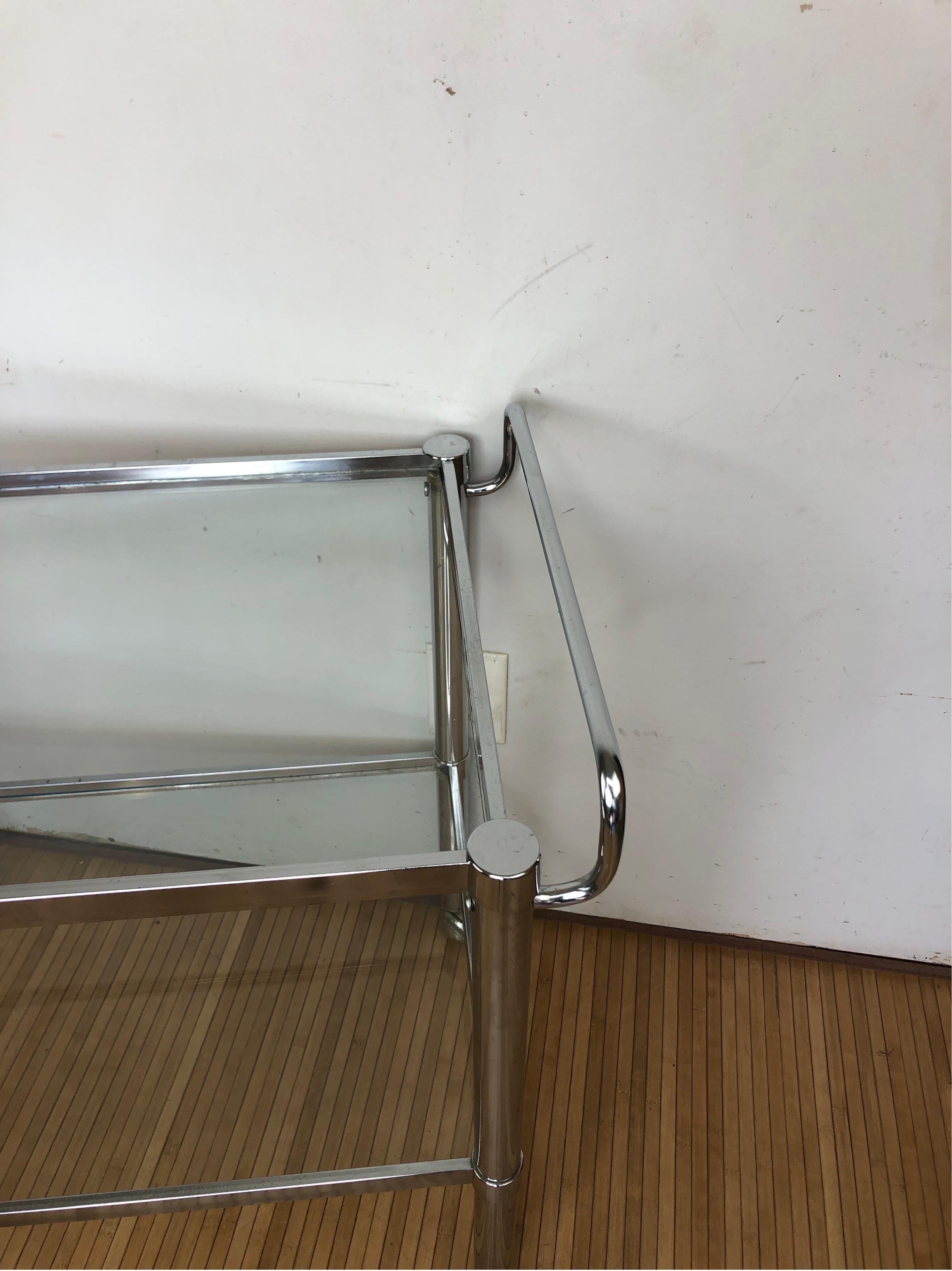This vintage mid-century glass and chrome bar cart is a stylish and functional piece of furniture that would be a great addition to any home. The cart is made of clear glass and chrome, and has a simple yet elegant design. It has two shelves, one