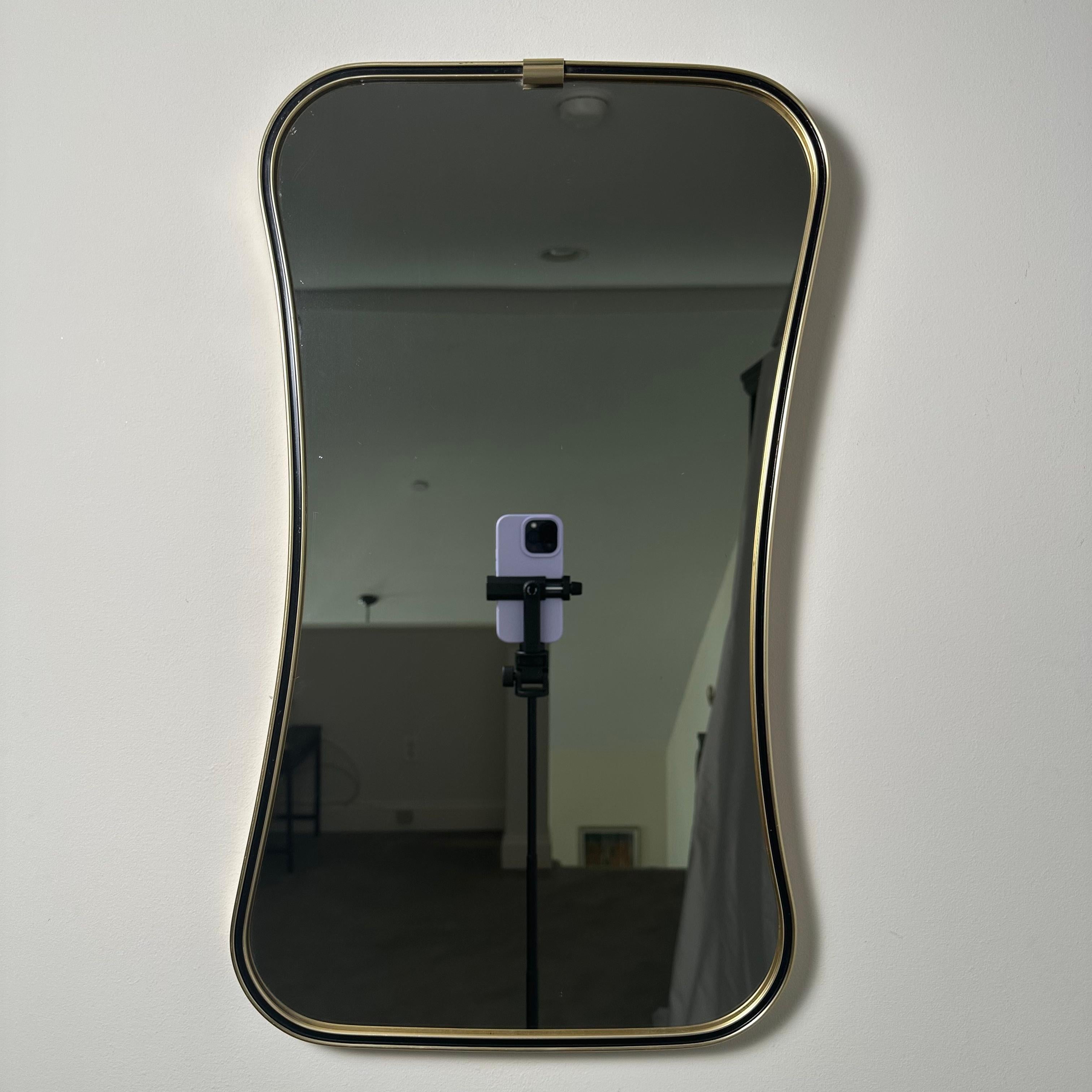 Vintage Italian Mid-Century Modern hanging wall mirror framed in brass with black enamel or lacquer border surround. Featuring a gorgeous and unique asymmetrical near-hourglass shape. While looking at first glance like an hourglass shape, the curves