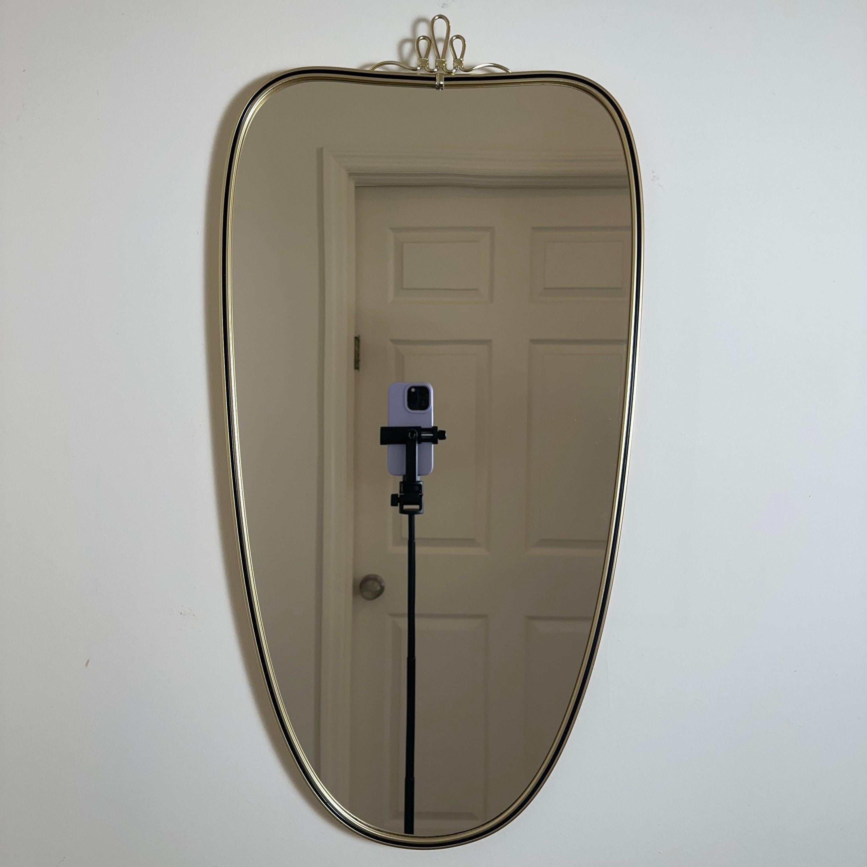 Vintage mid-century Italian brass wall mirror. In the shape of a subtle shield or heart, ringed by a black border. Topped with a looping ribbon detail, a wavy squiggle motif inspired by the designer Josef Frank. 

Right in size for a bathroom vanity