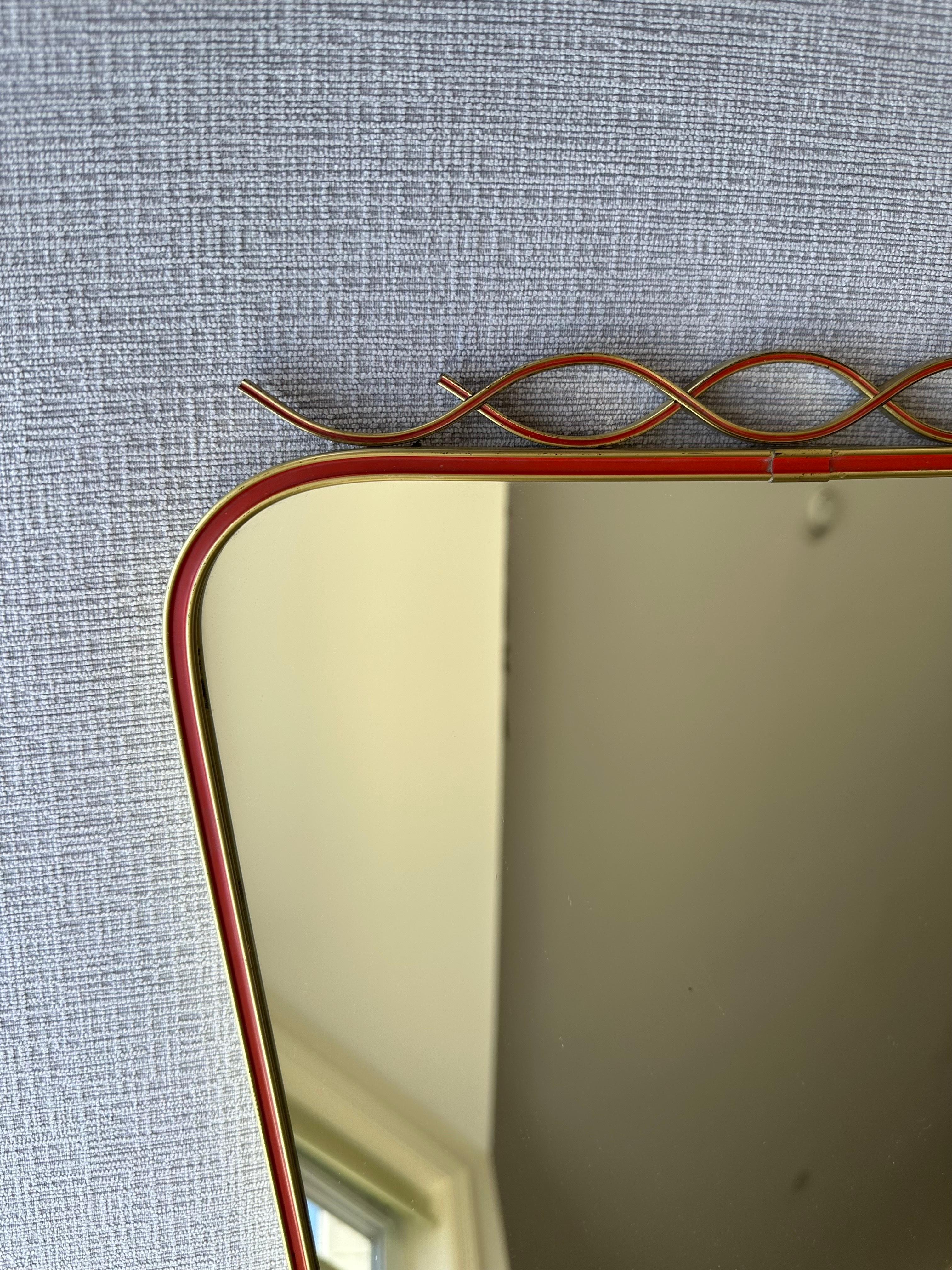Vintage Italian Mid-Century Modern brass wall mirror. Featuring an asymmetrical shield shape that is widest at the top. This hanging mirror is crafted in a brass frame with a red lacquer detail surround. At the top, an intersecting wave pattern