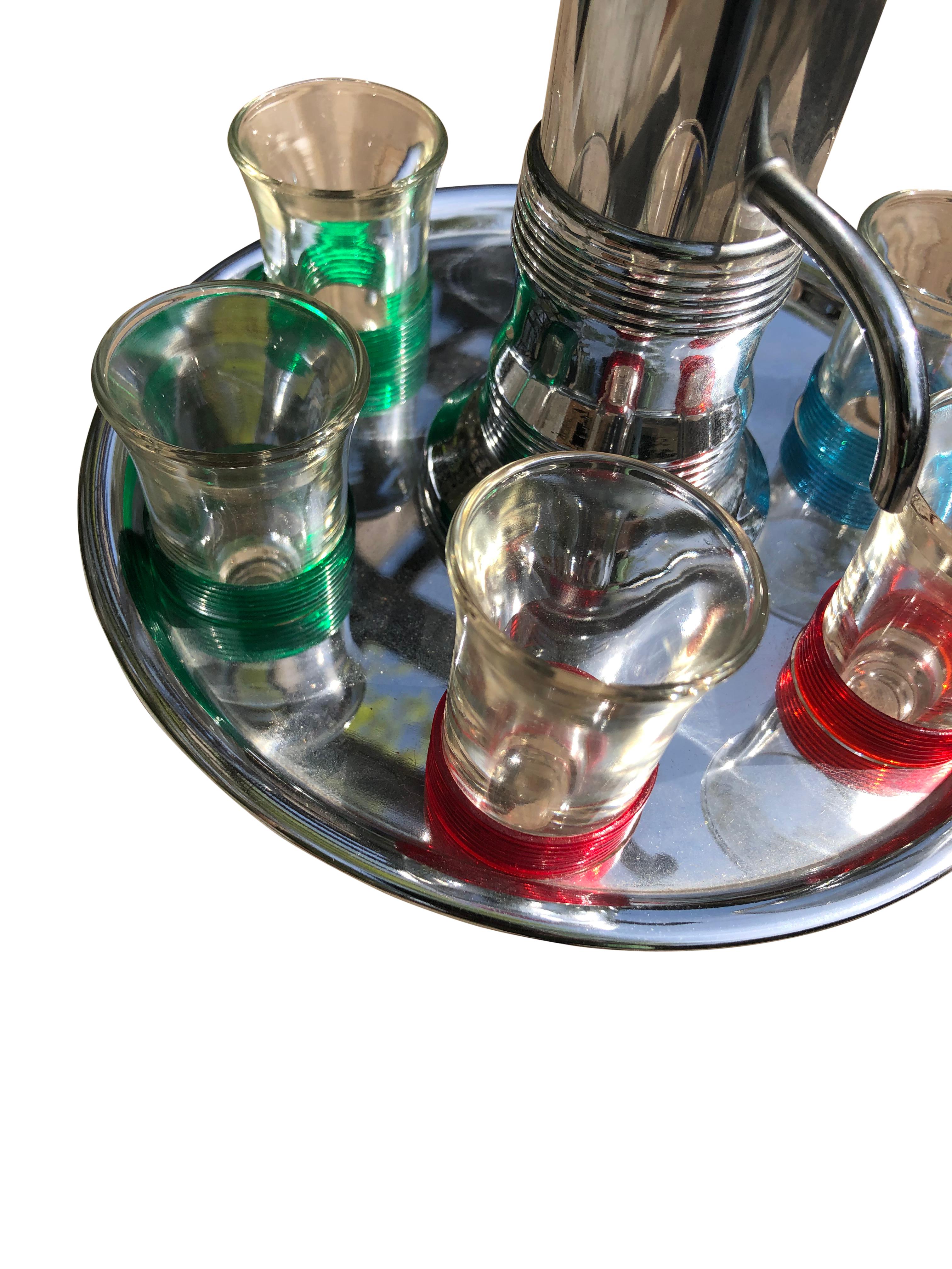 Vintage Mid-Century Carousel Liquor Dispenser with Set of 6 Shot Glasses sitting on a circular chrome plate with 6 cut-out slots to hold the shot glasses. Shot glasses are grouped in pairs with matching colored bands. There are two glasses with