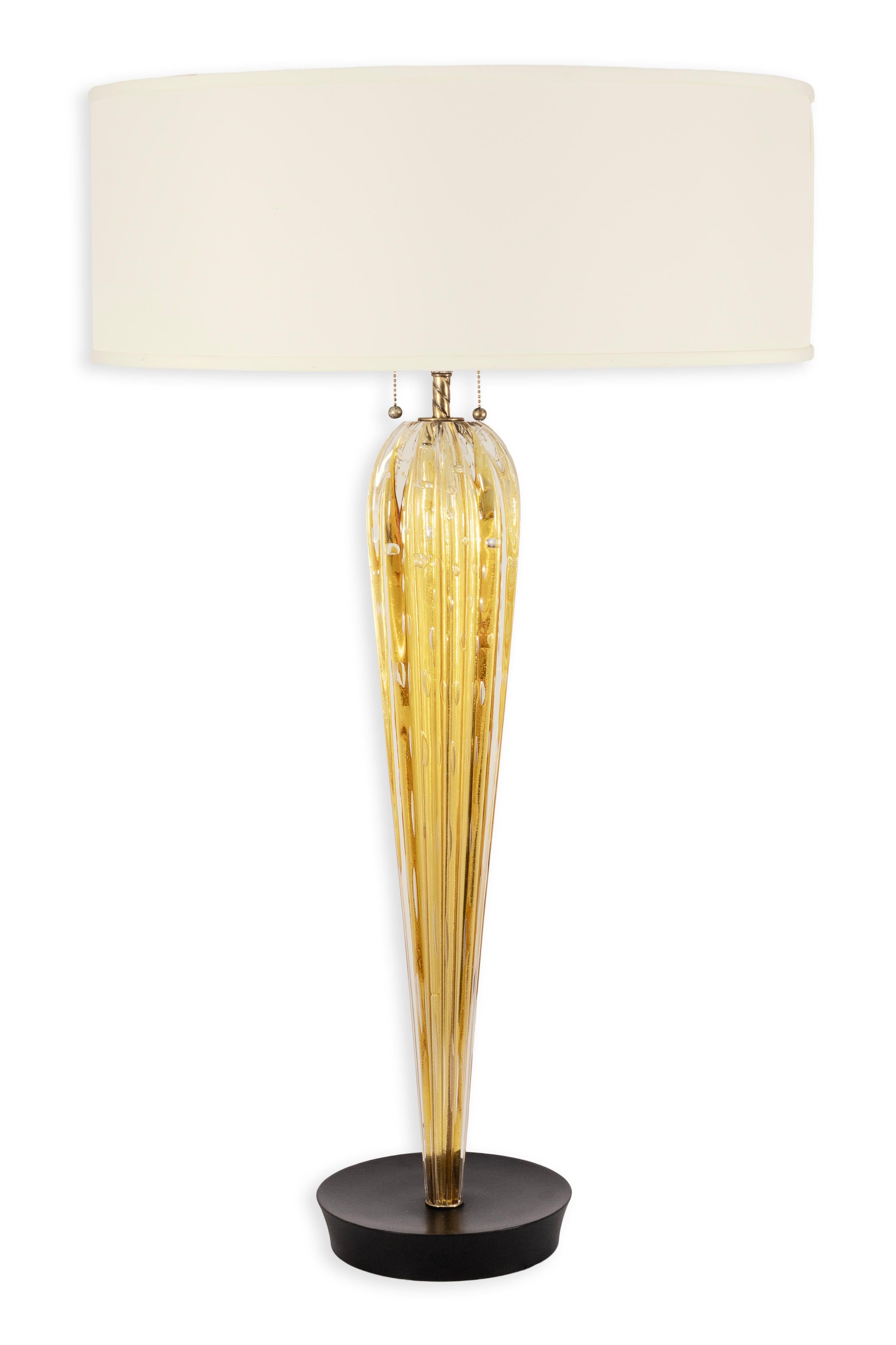 Diese elegante Tischleuchte verfügt über einen hohen, länglichen Glassockel in einem satten, goldenen Bernsteinton mit klaren, gerippten Details, die das Licht schön einfangen und reflektieren. Die skulpturale Form verjüngt sich anmutig zu einem