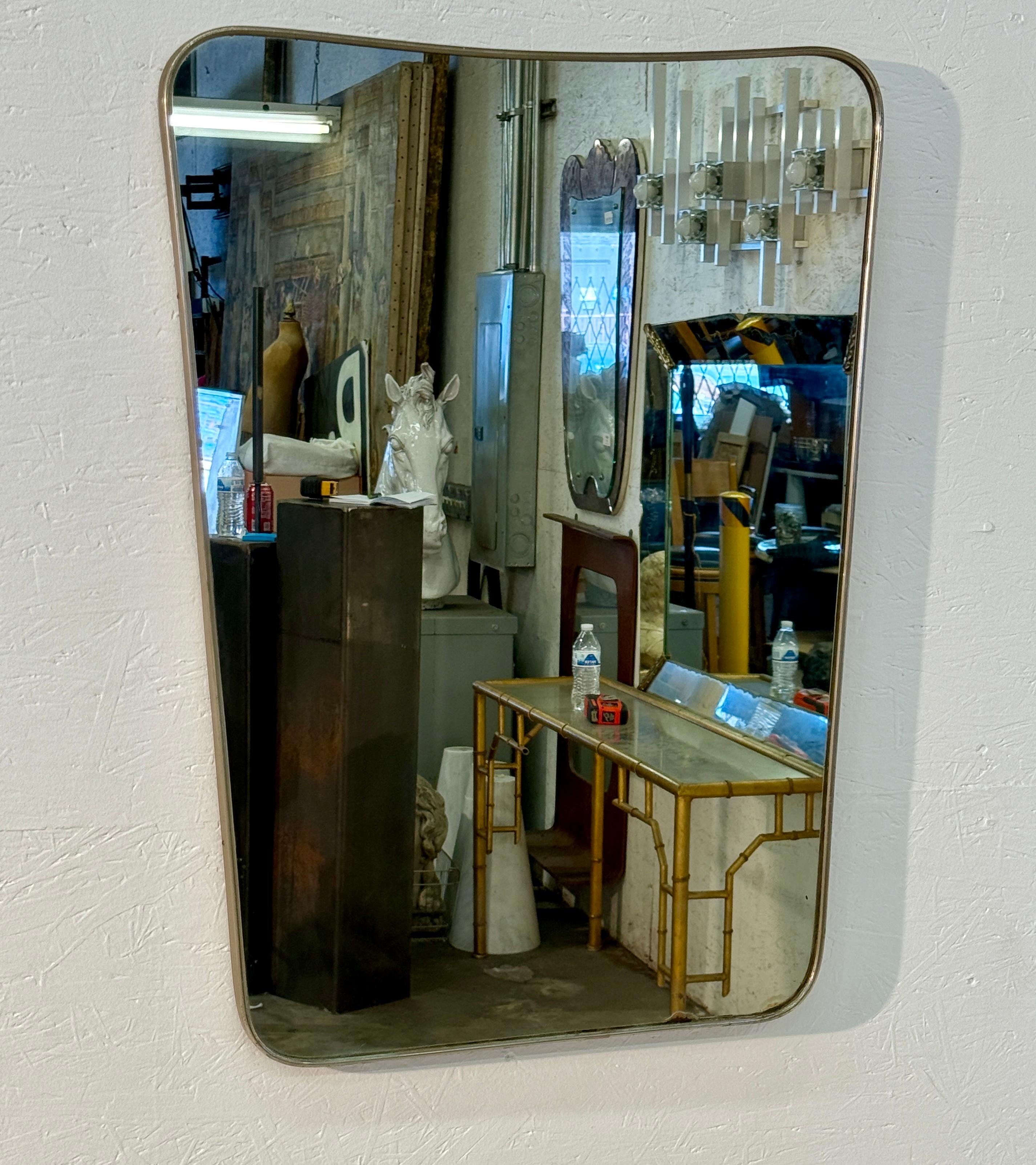 Espejo de pared moderno de mediados de siglo con forma rectangular asimétrica y bordes sutilmente curvados, que le dan un aspecto elegante y orgánico. El espejo está enmarcado en un fino marco de metal cepillado, que ha adquirido una pátina