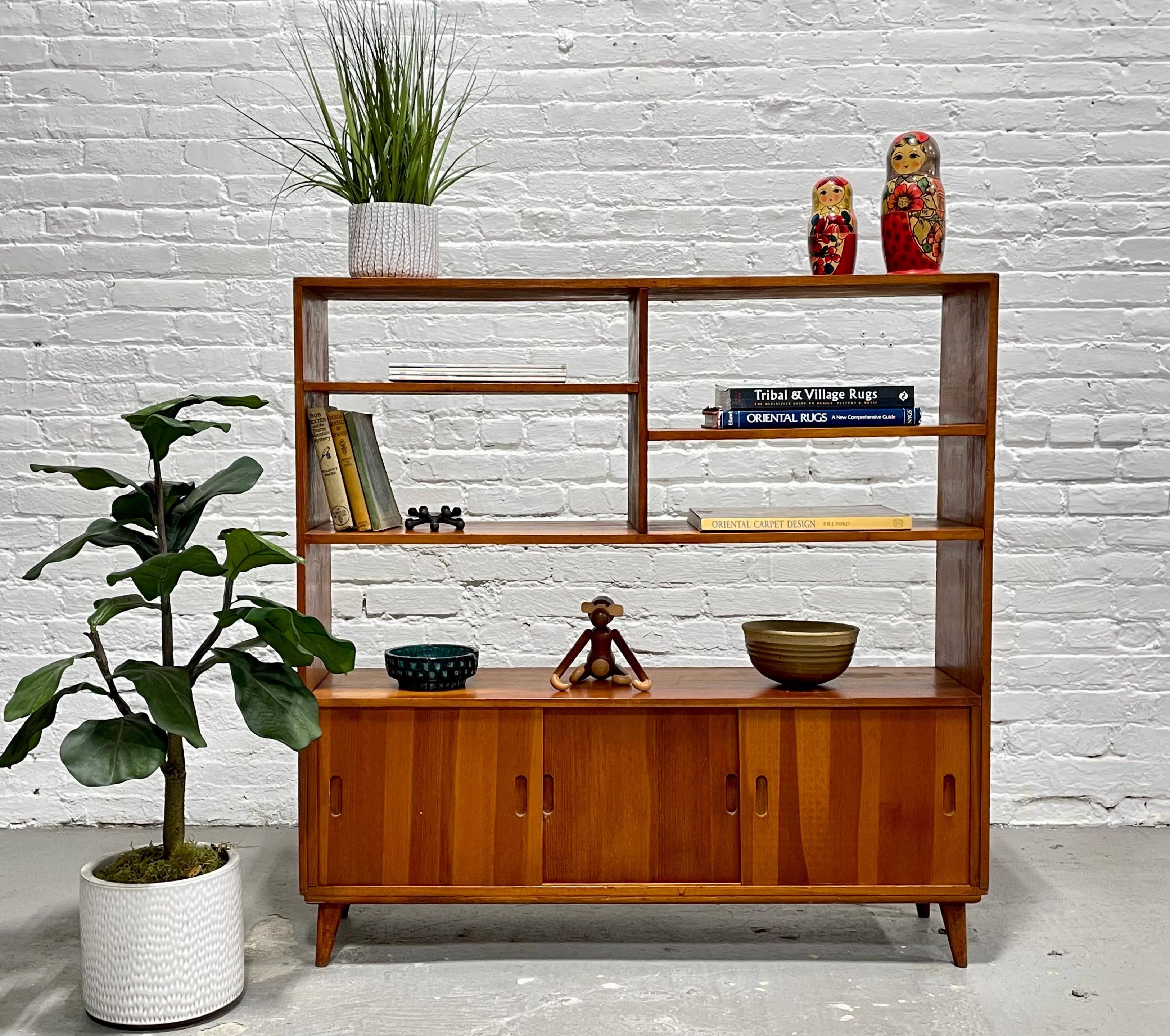 Mid-Century Modern bookcase / space divider featuring a thoughtful combination of shelving and a hidden cubby area behind three sliding doors. This handsome bookcase offers a variety of shelving to display your books or collectables and a lower