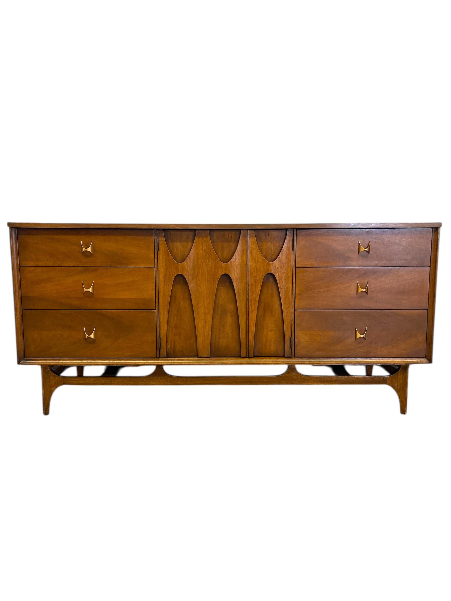 Esta cómoda vintage de 9 cajones es una pieza icónica de la colección Broyhill Brasilia, célebre por sus líneas escultóricas y su inspiración arquitectónica. Fabricada en nogal macizo, la cómoda luce el característico detalle arqueado en forma de