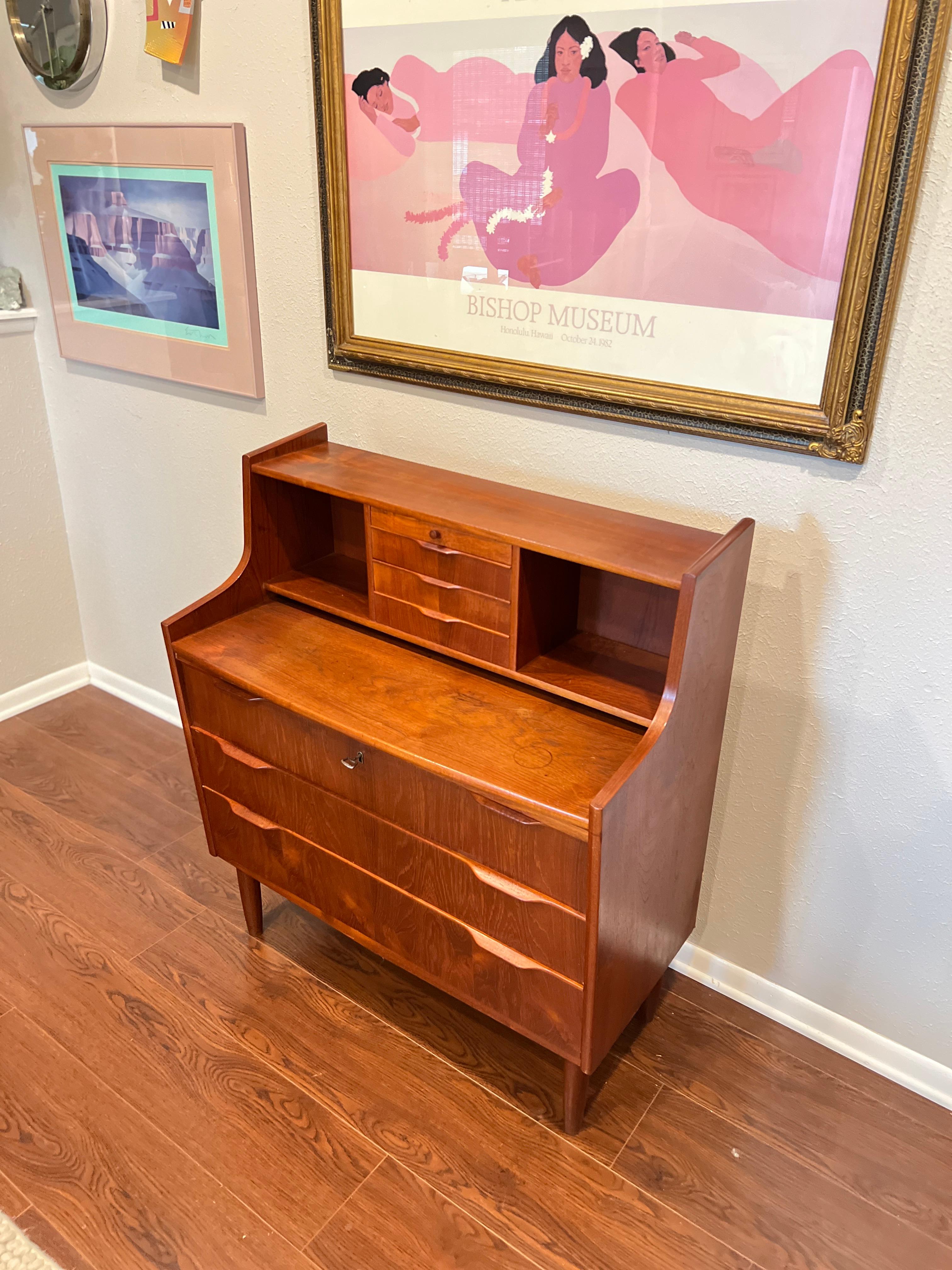 Danish style midcentury secretary desk from the 1960s, made of teak veneer. Top section has one sliding tray above 3 stacked drawers at center, flanked by two open storage niches. Also has a sliding leaf desk top and underneath are three bottom