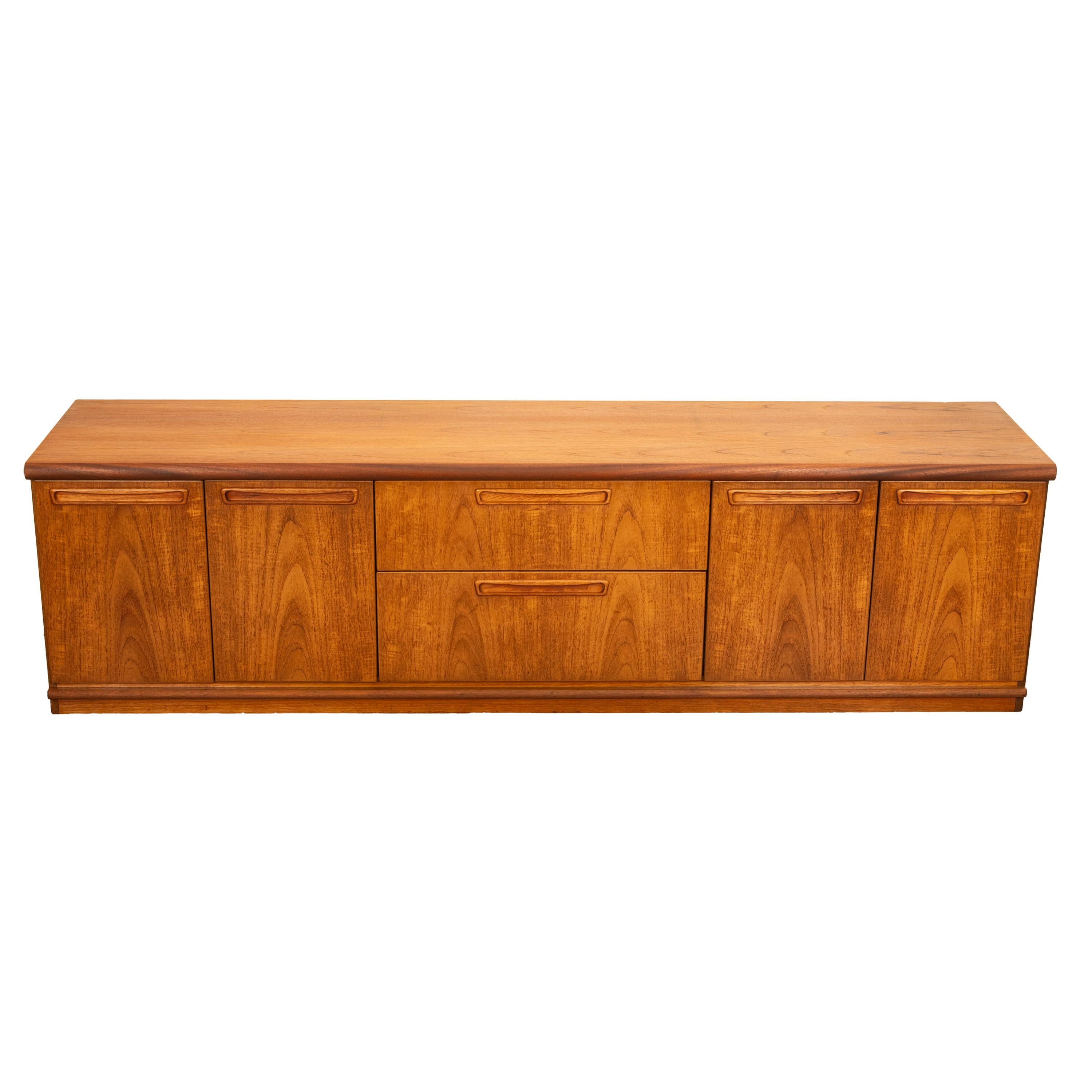 A good long original vintage Mid Century Modern Danish style teak credenza, by Avalon, 1960s.
This long low credenza made from teak has twin cupboard doors at each end, the one on the left side enclosing a drawer and a shelf, to the center is a pair