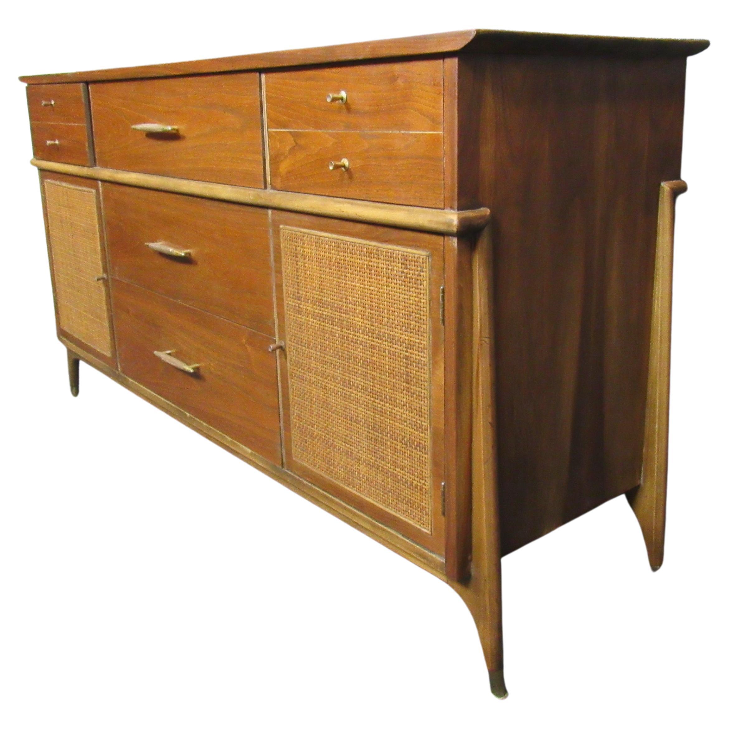 Amazing vintage mid-century dresser. The marvelous wood grain shines through with beautiful carved binding and contrasting wicker and brass accents. Features seven pull-out drawers with two large side cabinets for plenty of storage and