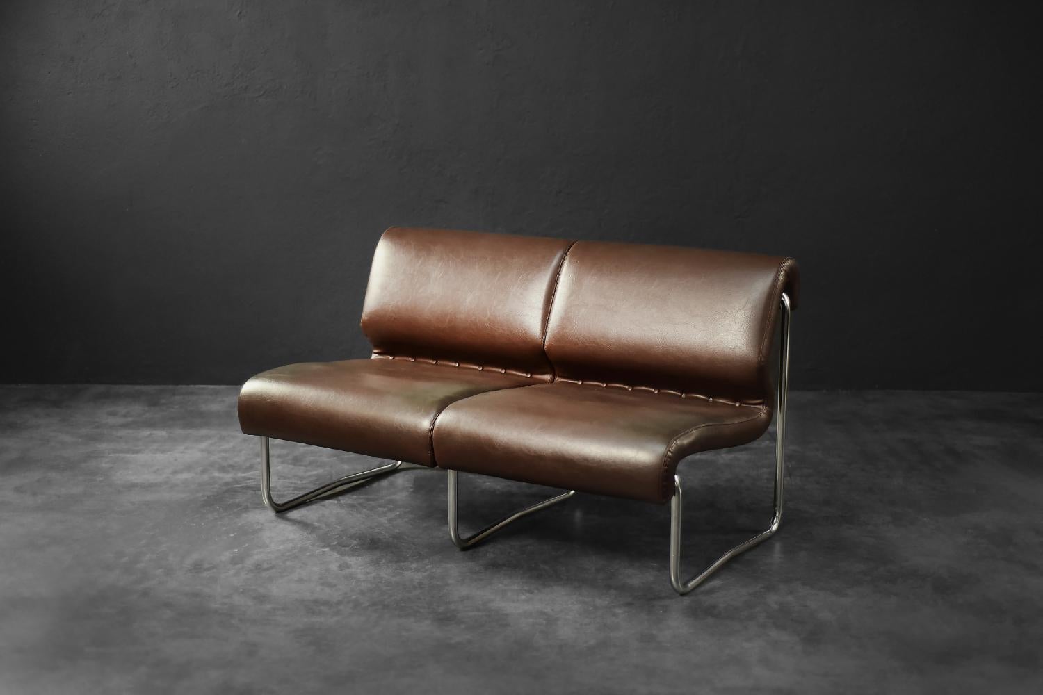 This two-seater sofa was probably manufactured in Germany during the 1970s. This piece is inspired by the minimalist aesthetic of the Bauhaus school with a 1970s modernist twist. It is upholstered in brown synthetic leather with a visible patina,
