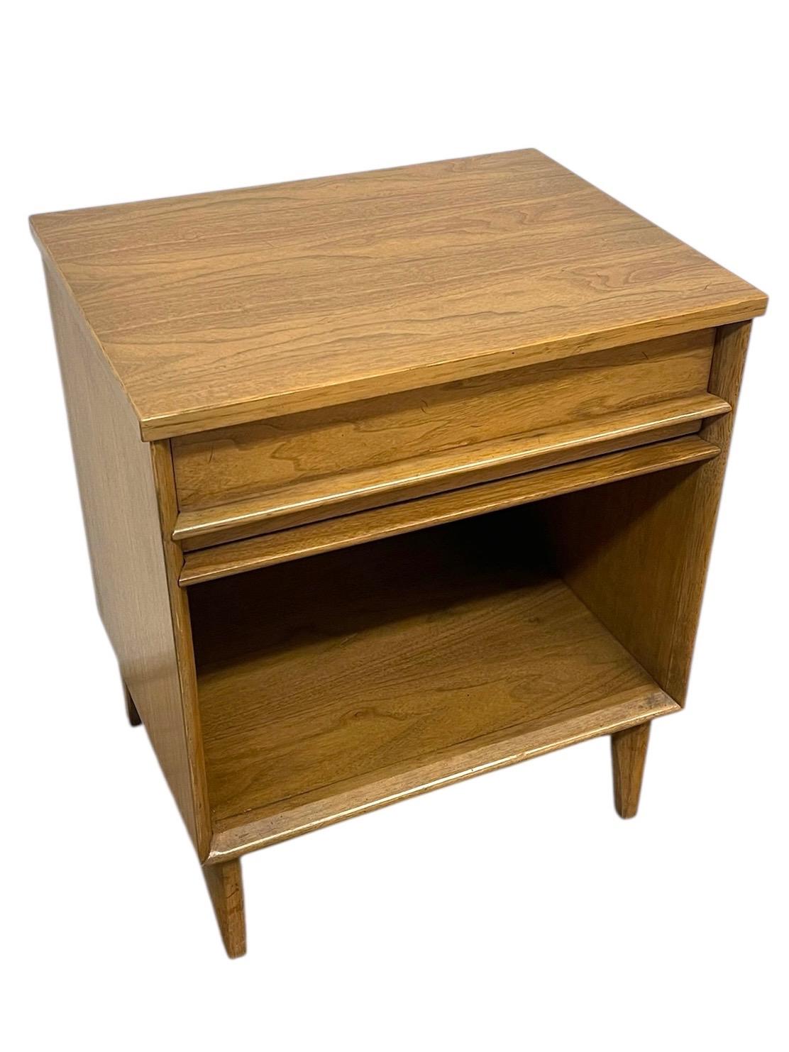  This vintage mid-century modern nightstand features clean lines, tapered legs, and a warm blonde finish. The design includes a single pull-out drawer with integrated molded wood pulls and an open lower shelf, offering both storage and display