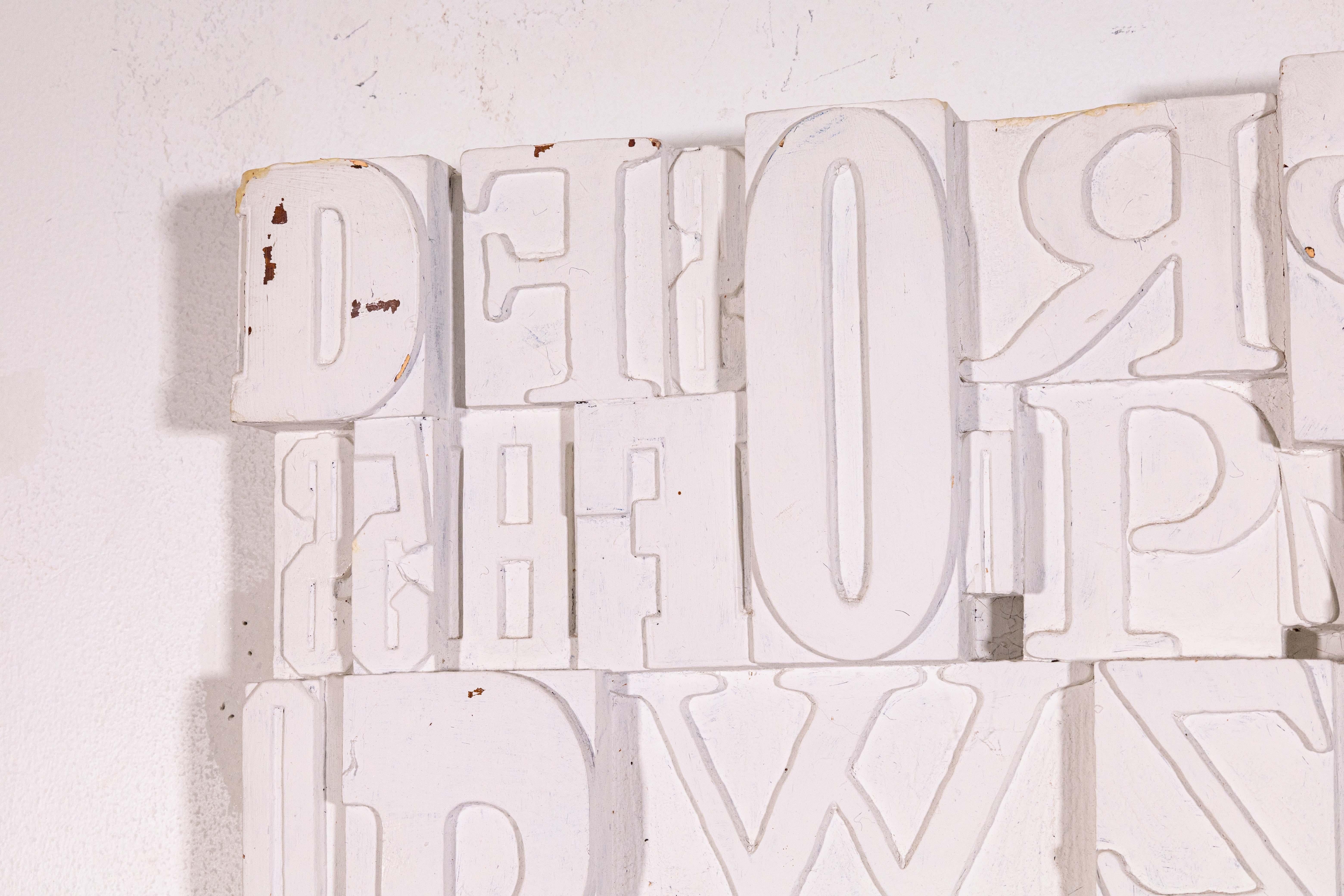 A unique mid century modern wall hanging sculpture composed of white painted typeface letters. Circa 1960s. A minimal aesthetic yet also provides a sense of nostalgia with the display of vintage printing methods. The composition is inspired by