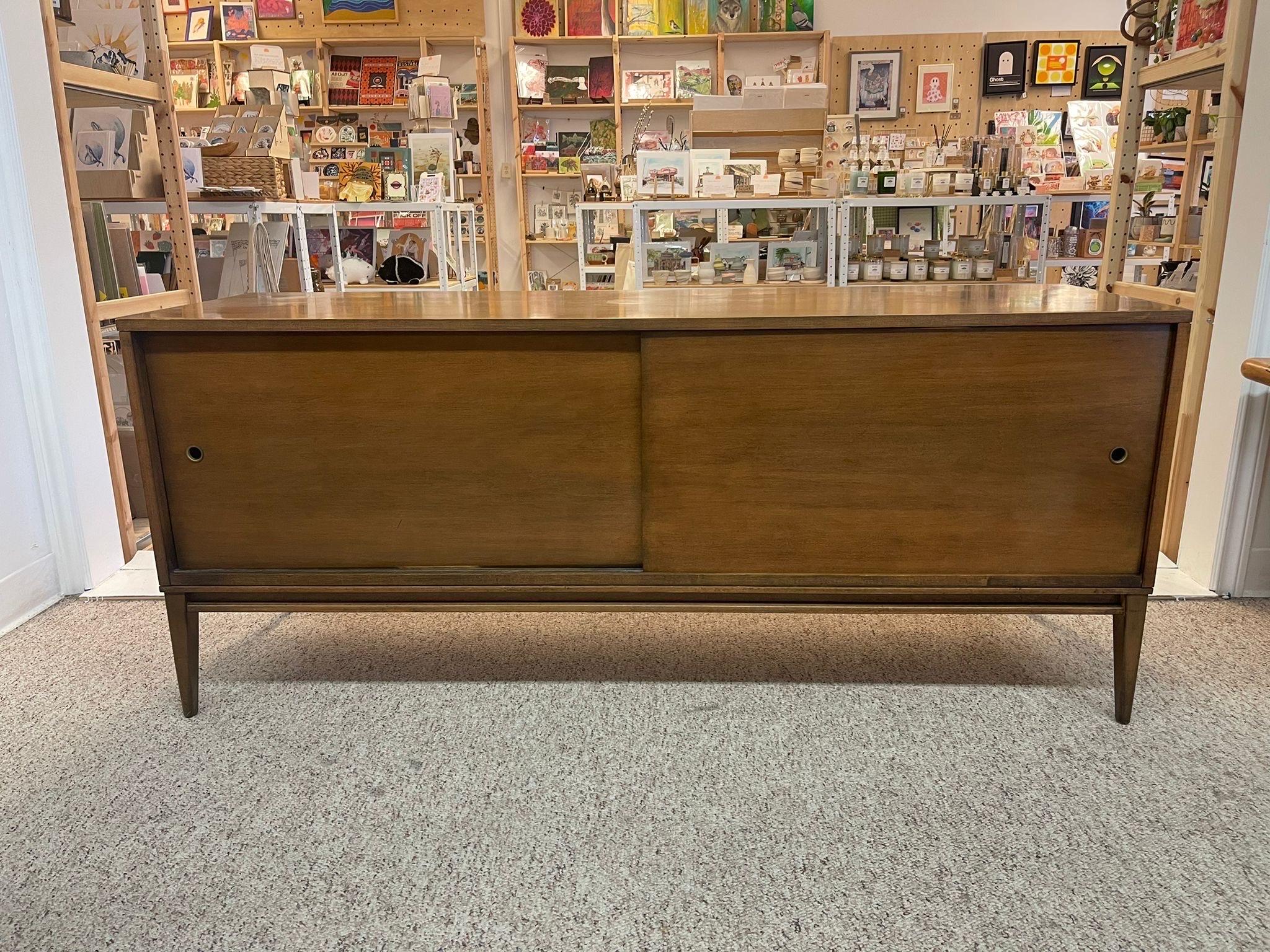 Each side of the cabinet has adjustable shelving. Tapered Legs. Unique handles to the sliding doors, a circular hole with metal ( brass toned ) lining. Circa 1950s. By Paul McCobb for Winchendon Furniture. Vintage Condition Consistent with Age as