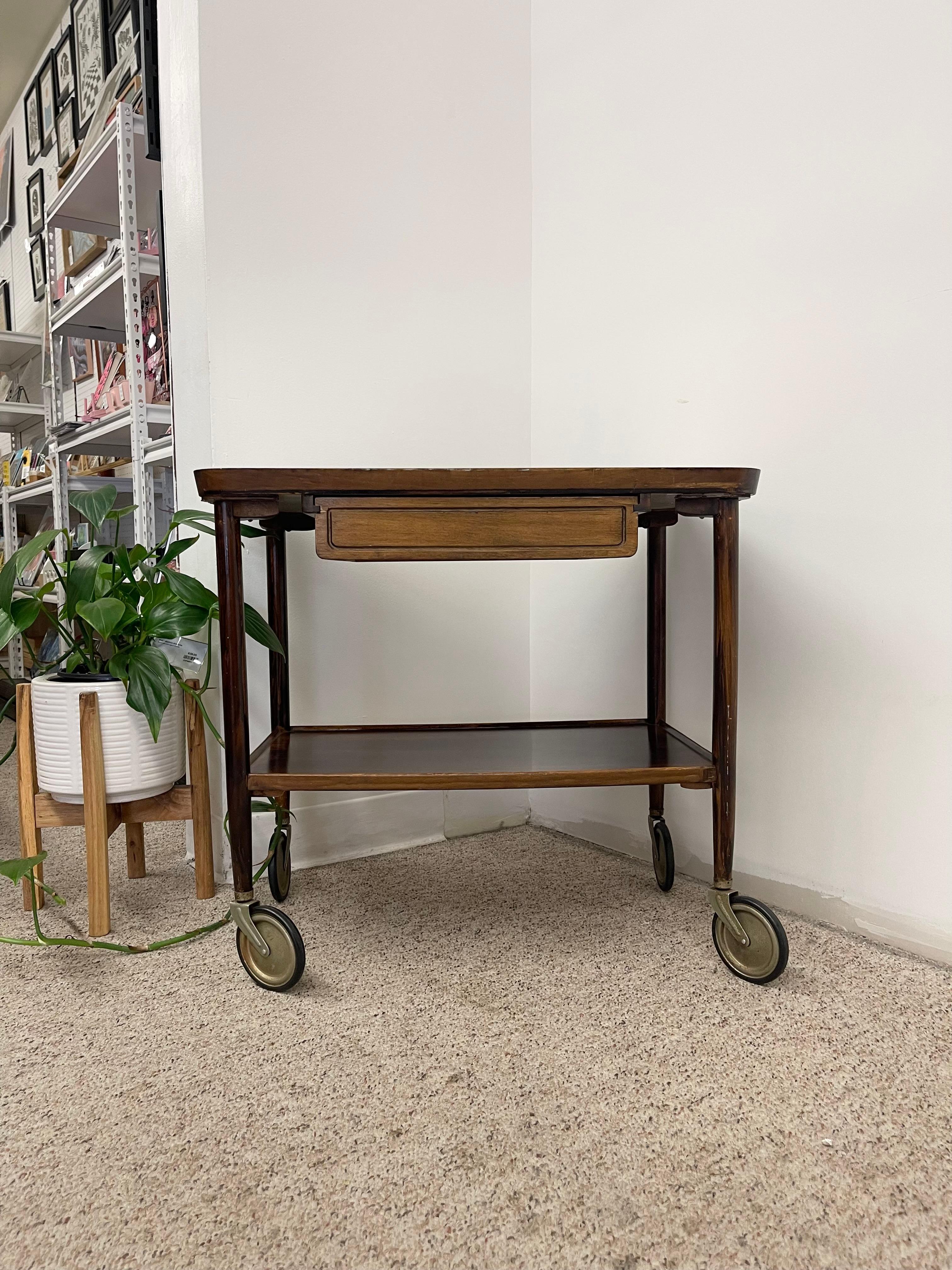 This vintage mid-century modern rolling bar cart combines practicality with timeless design. Crafted with a warm wood frame, it features a rectangular top inset with a white textured surface, framed by raised wooden edges for both style and