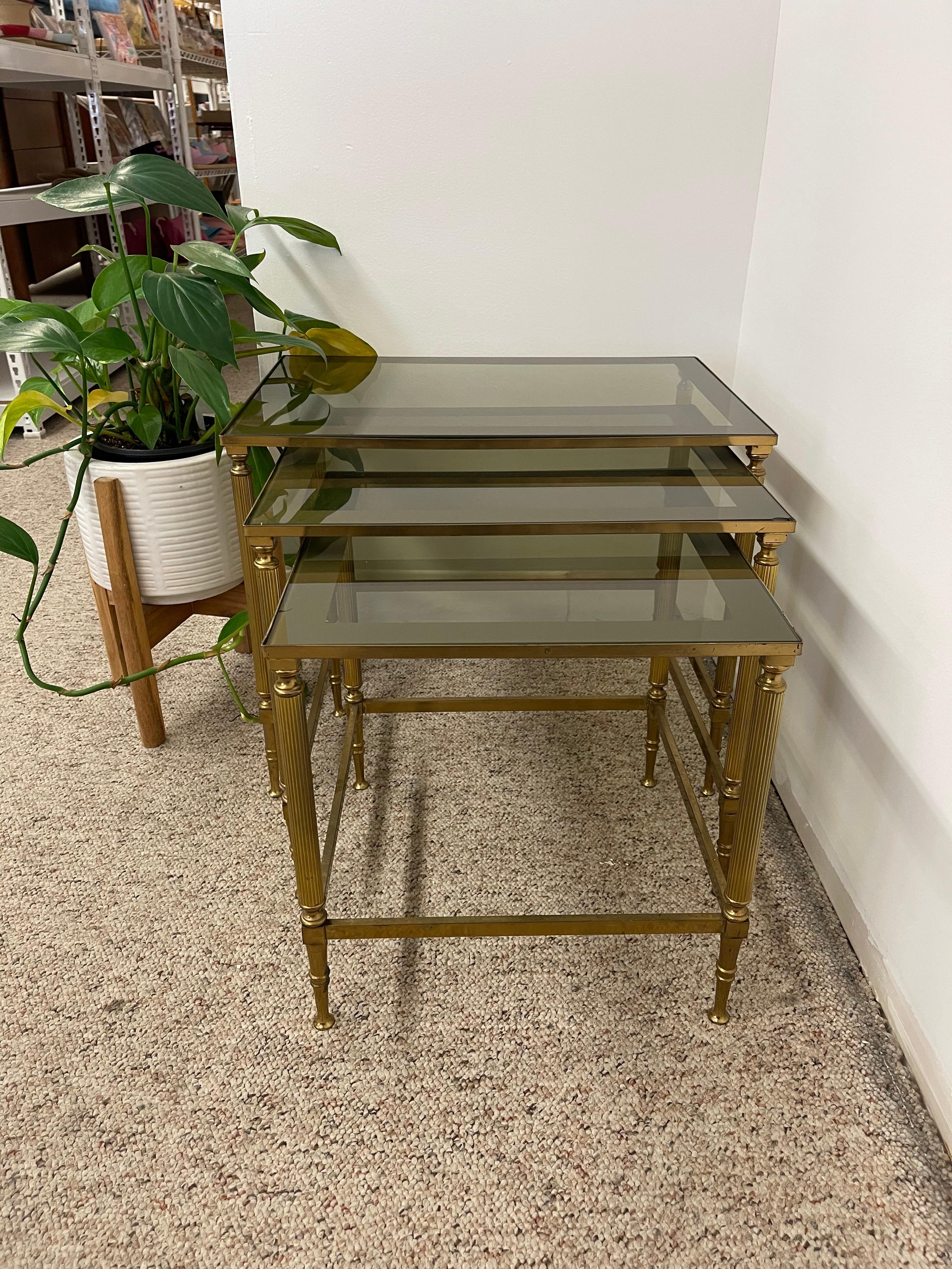  Elegant set of three mid-century modern nesting tables featuring smoked glass tops. Each table is designed with reeded, fluted legs and finely detailed accents, giving the set a sophisticated, neoclassical-inspired aesthetic. The smoked glass