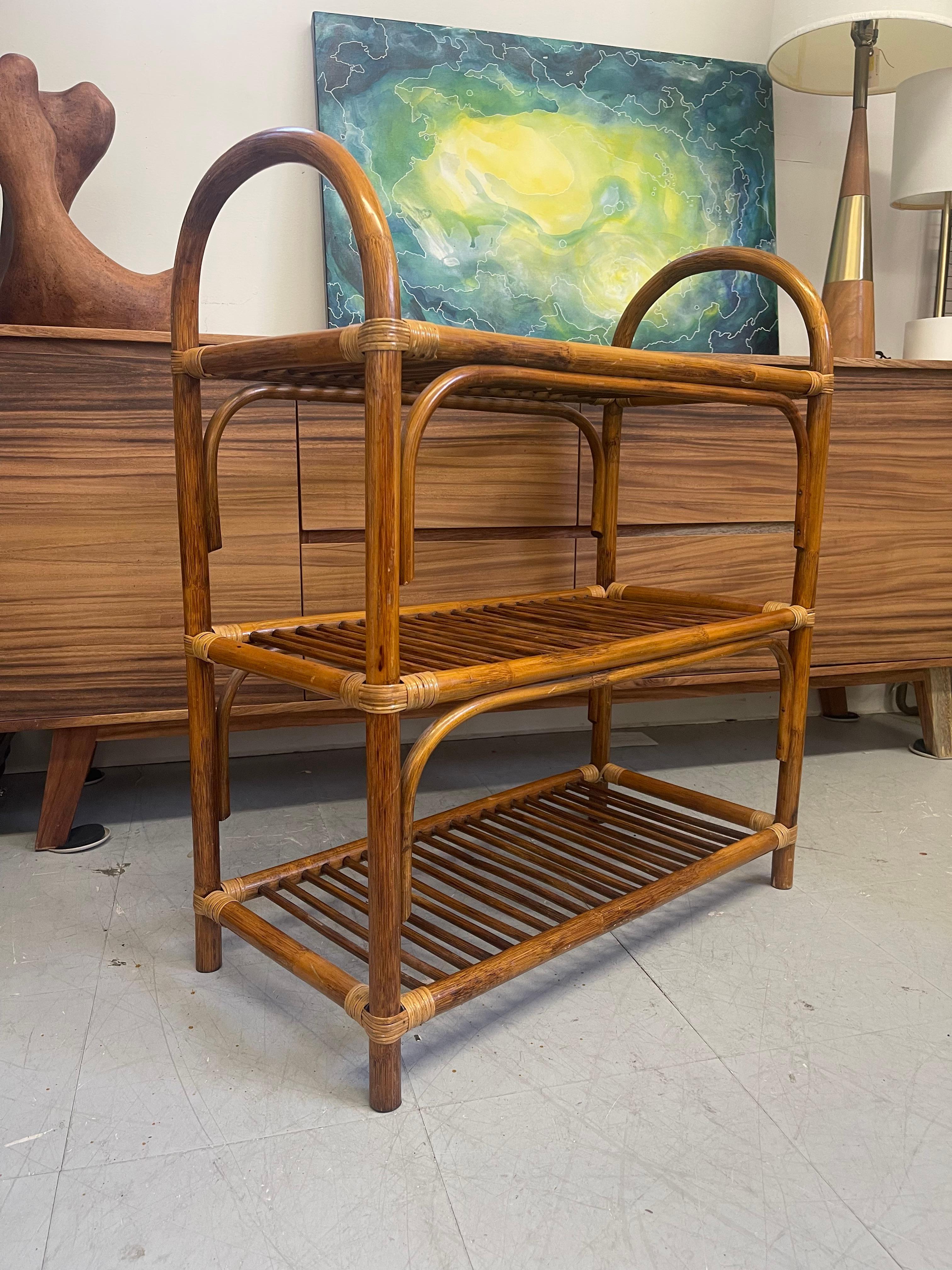 Scaffale vintage a tre livelli in rattan di bambù con maniglie laterali ad arco e scaffali aperti a doghe. Probabilmente è stato realizzato in bambù curvato con giunture avvolte in canne. La superficie presenta una patina calda e naturale, con