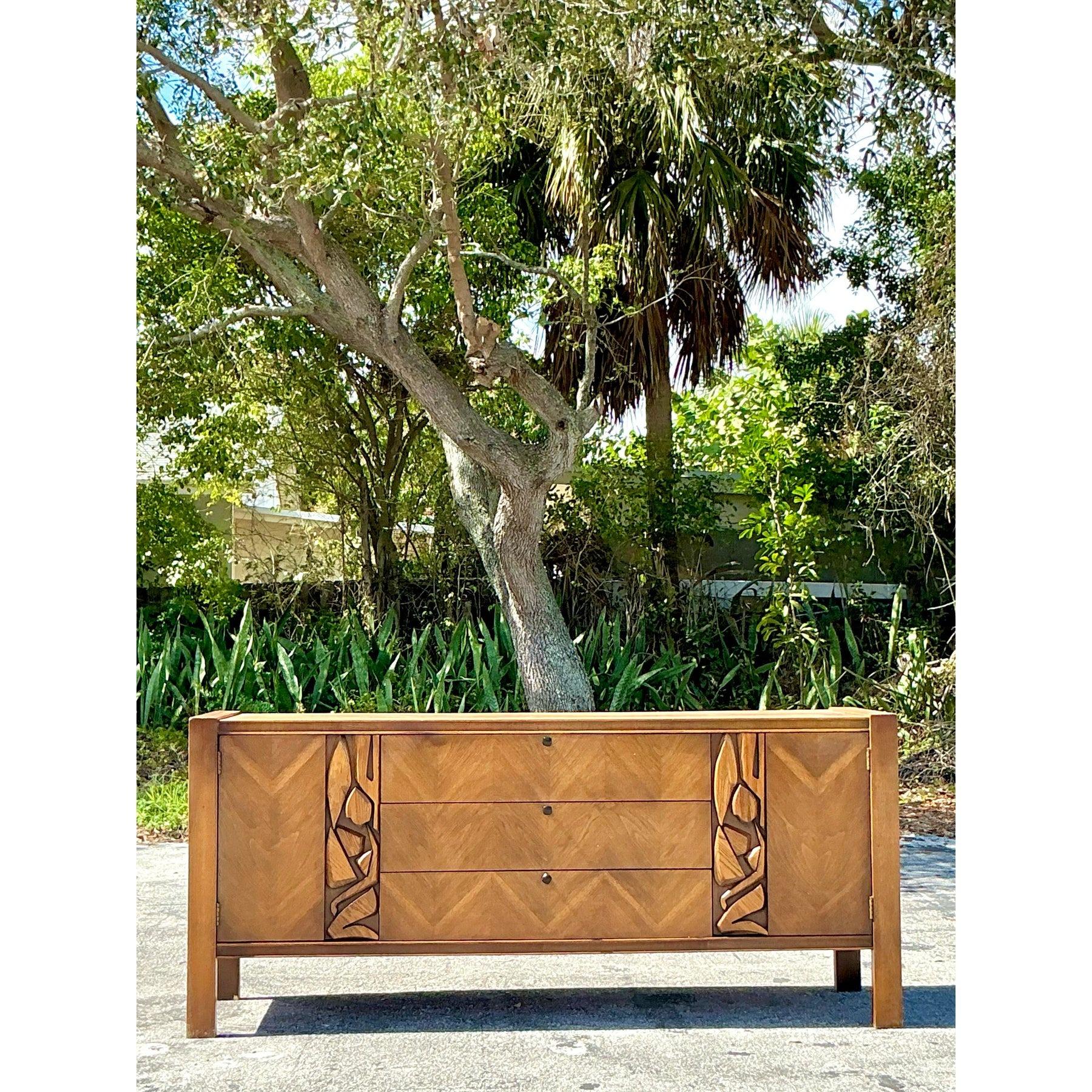 Haz una declaración audaz con este impresionante aparador moderno de mediados de siglo de United Furniture, que presenta una sorprendente mezcla de tonos de madera natural y esculturales detalles de inspiración tiki. El llamativo patrón geométrico
