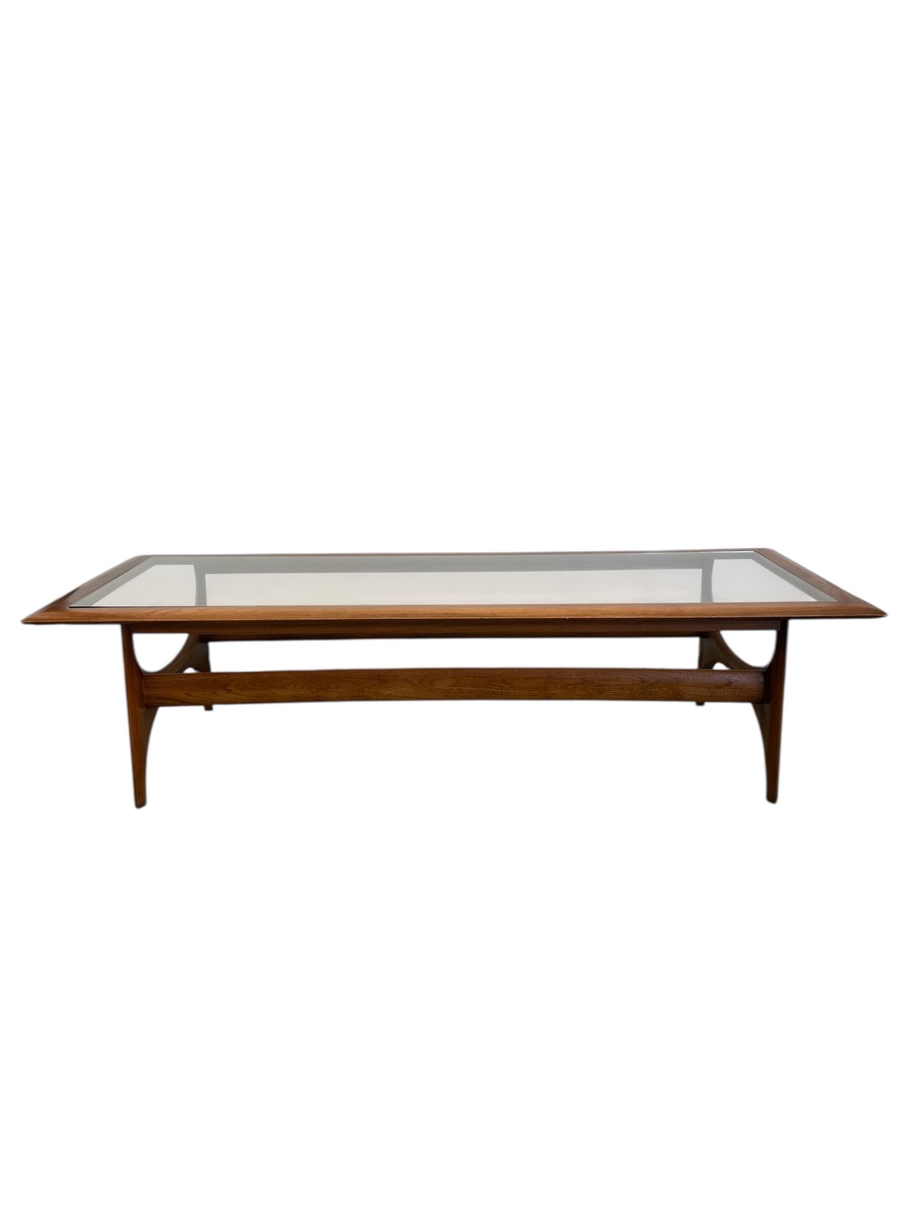 This vintage Mid-Century Modern coffee table by Lane Furniture exemplifies the sculptural elegance and refined craftsmanship of the era. The frame is constructed from walnut with beautifully contoured edges and arched stretchers that lend a light,