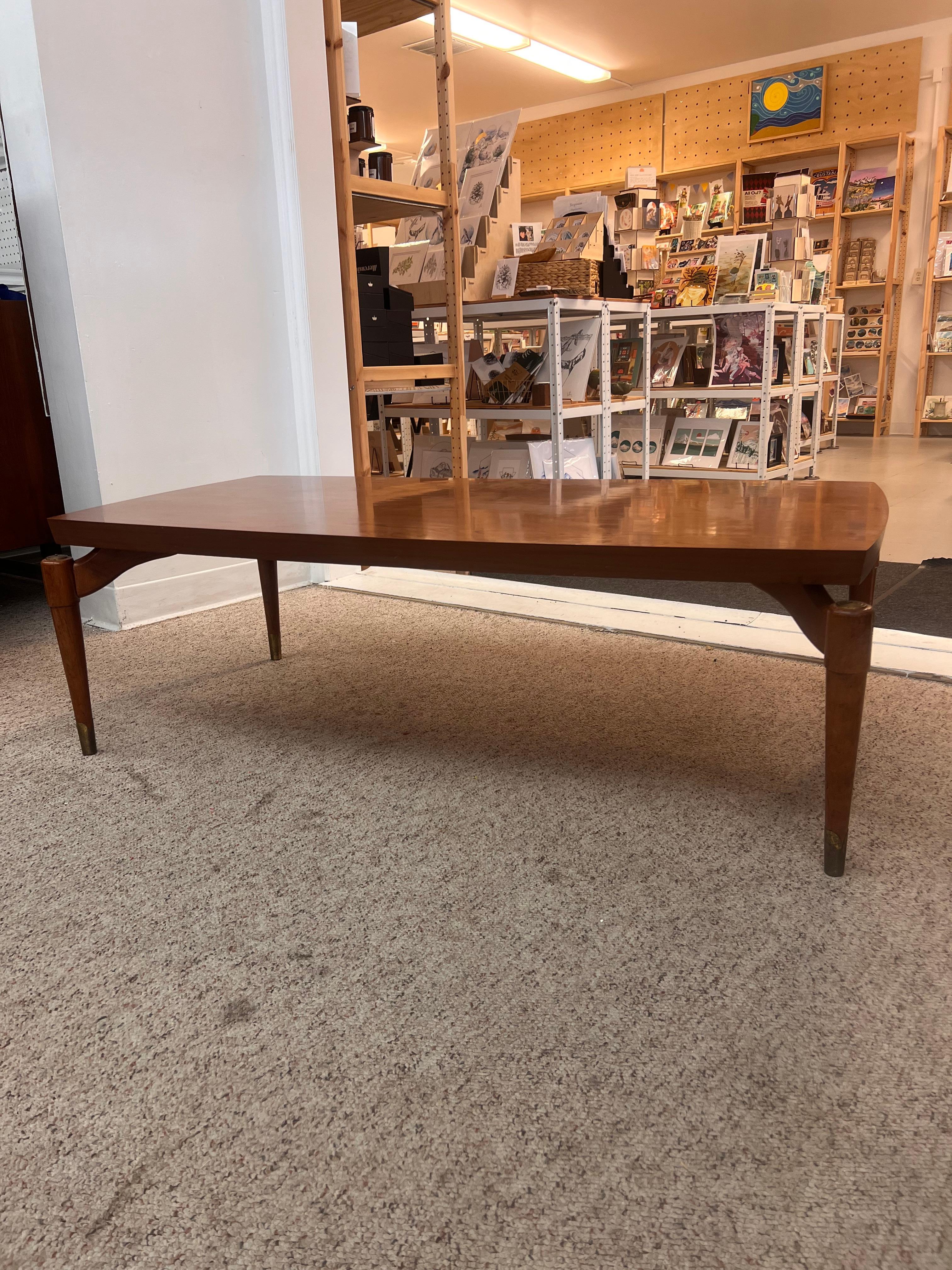 Cette table basse est une forme rectangulaire emblématique du milieu du siècle dernier. Il s'agit d'un très beau design avec un cadre en noyer, des pieds en forme de fusée et des pieds plafonnés en laiton. Le plateau est un faux noyer stratifié, ce